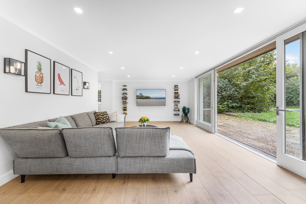 Modernes Wohnzimmer mit grauem Sofa, Holzboden und großen Fenstern mit Blick auf den Garten.
