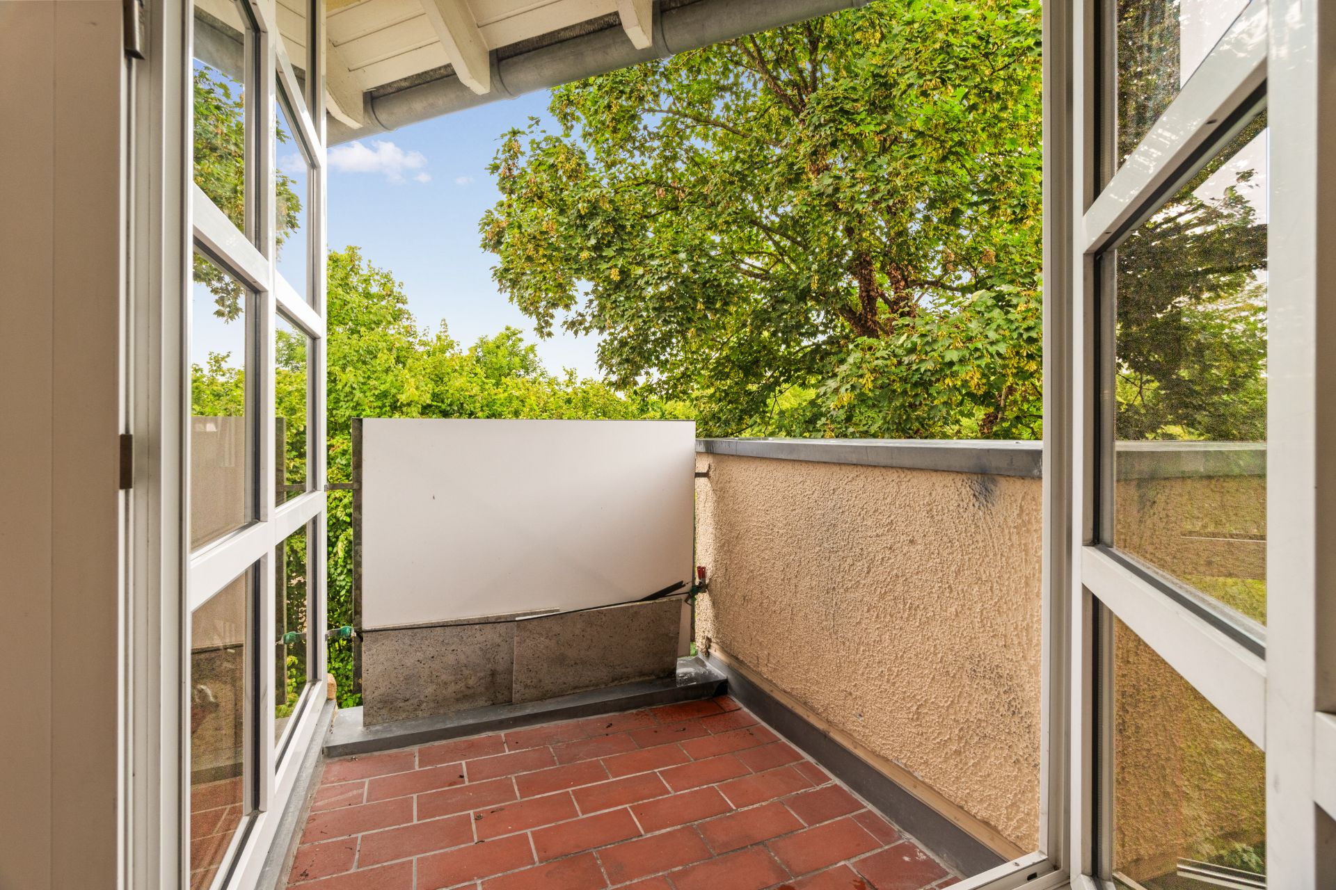 Kleiner Balkon mit roten Fliesen und Blick auf grüne Bäume.
