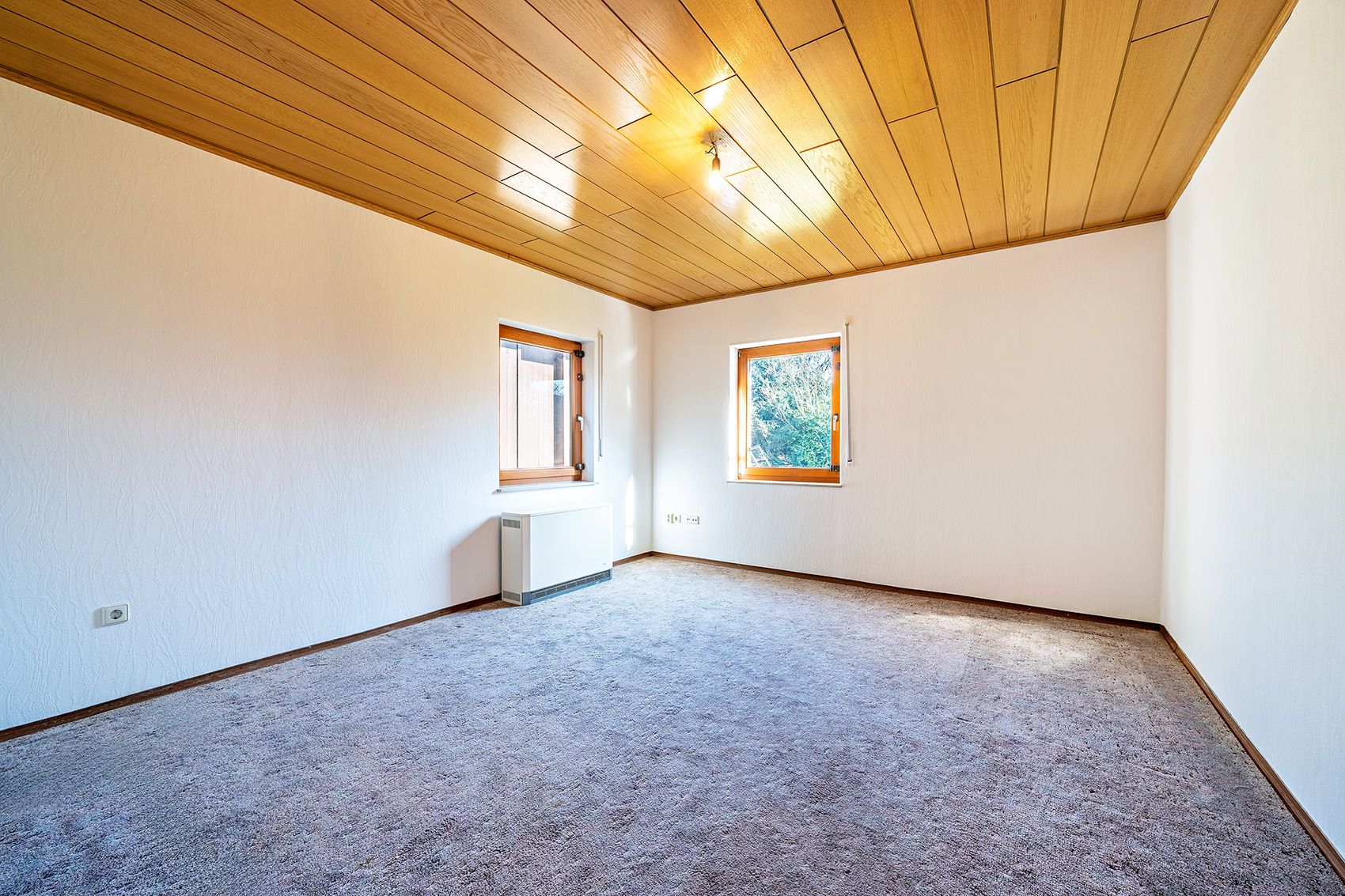 Leeres Zimmer mit Teppichboden, weißen Wänden, Holzdecke und zwei Fenstern.