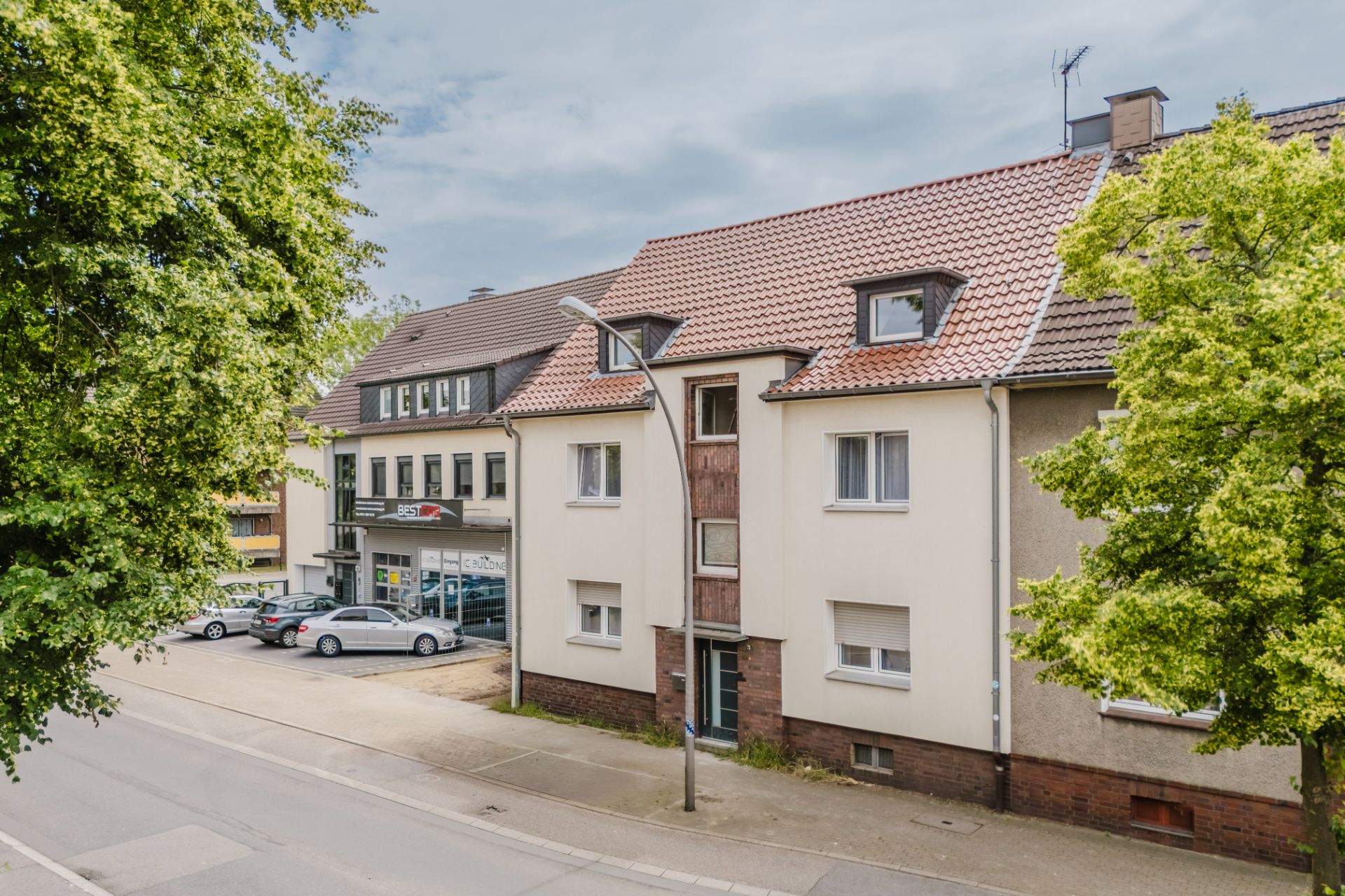Wohngebäude mit rotem Dach, mehreren Fenstern und Bäumen an einer ruhigen Straße.
