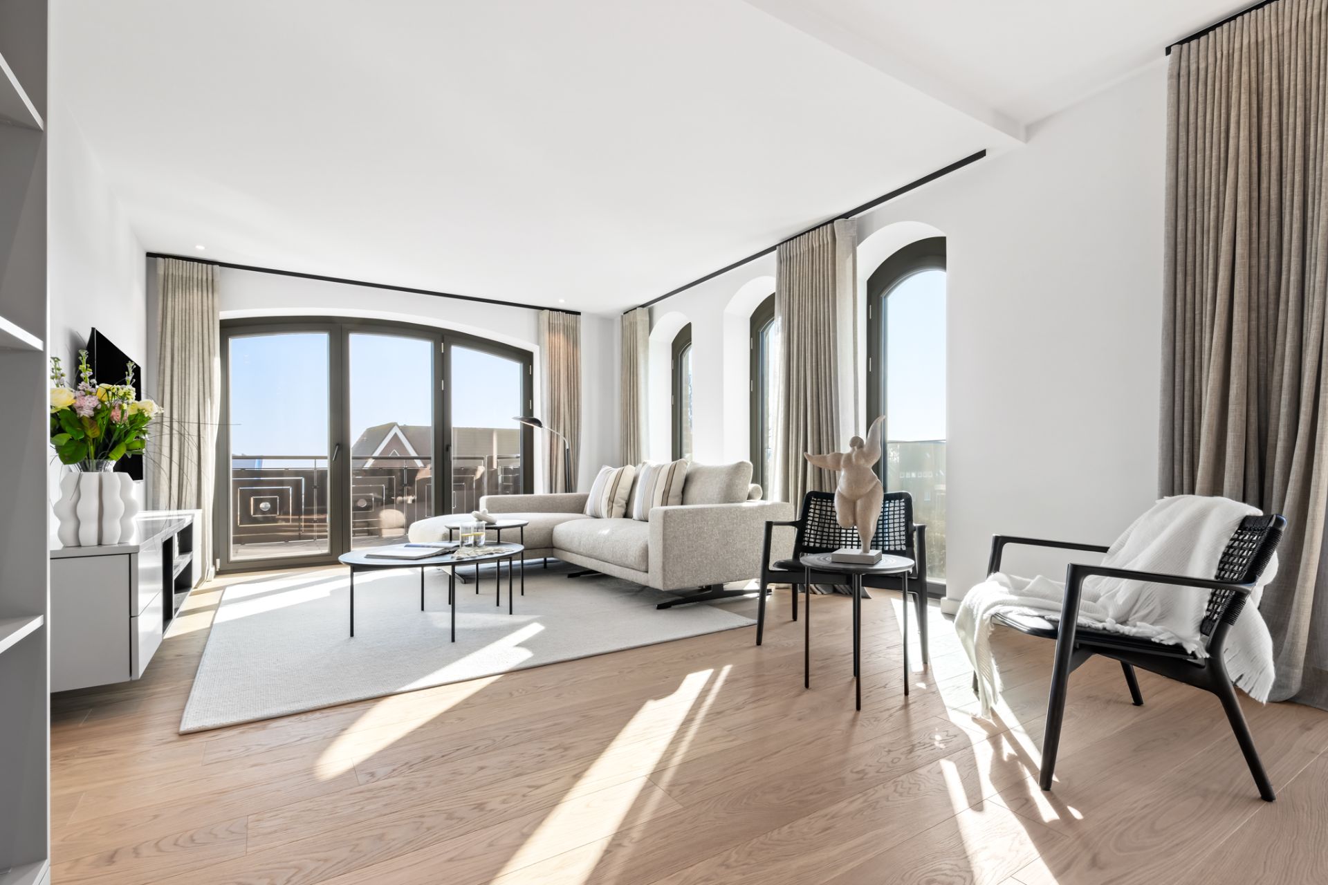 Helles Wohnzimmer mit Sofaecke, Sesseln, Couchtisch und großen Fenstern mit Vorhängen.