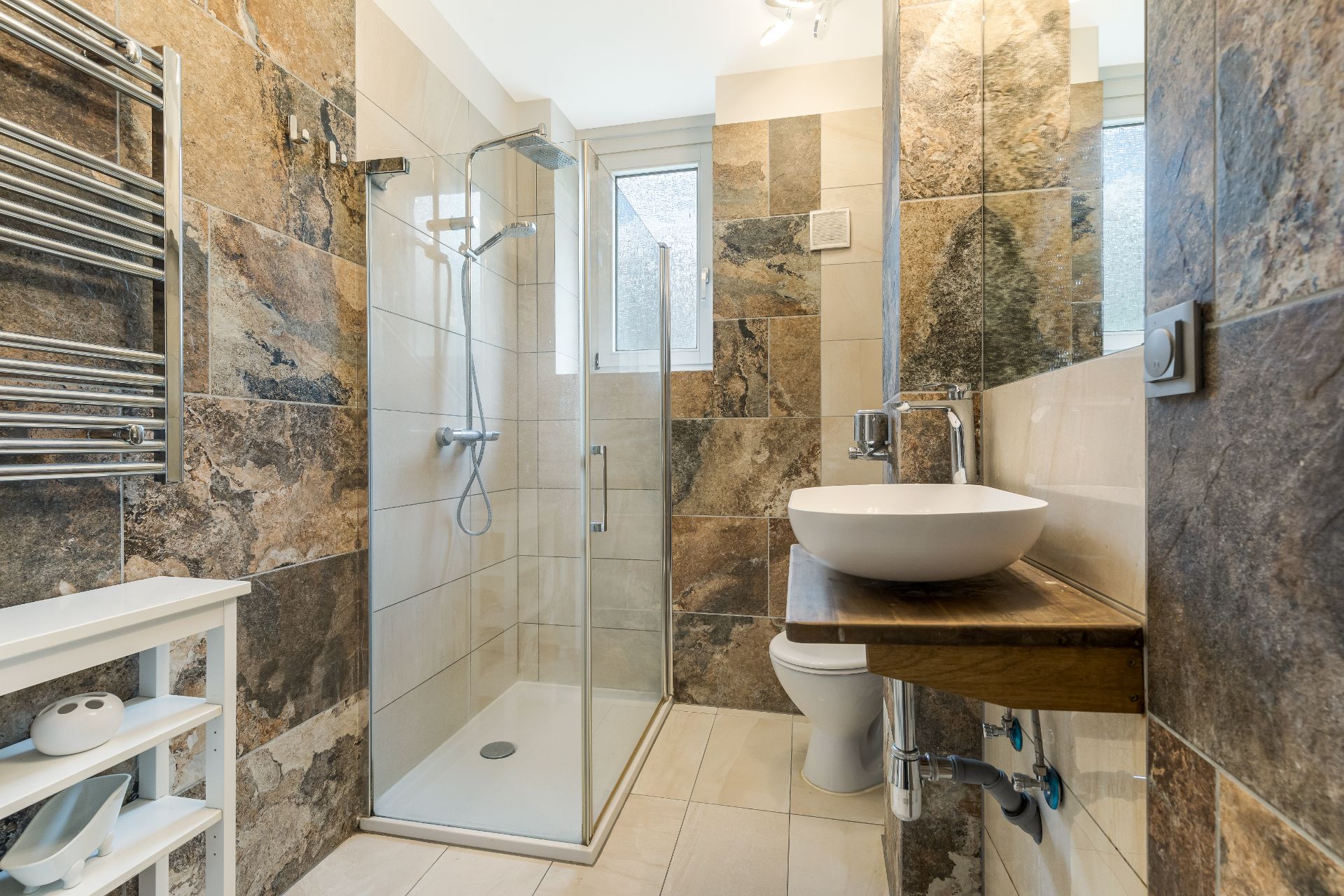 Modernes Badezimmer mit bodengleicher Dusche, Waschbecken und WC, gefliest mit Naturstein-Optik.