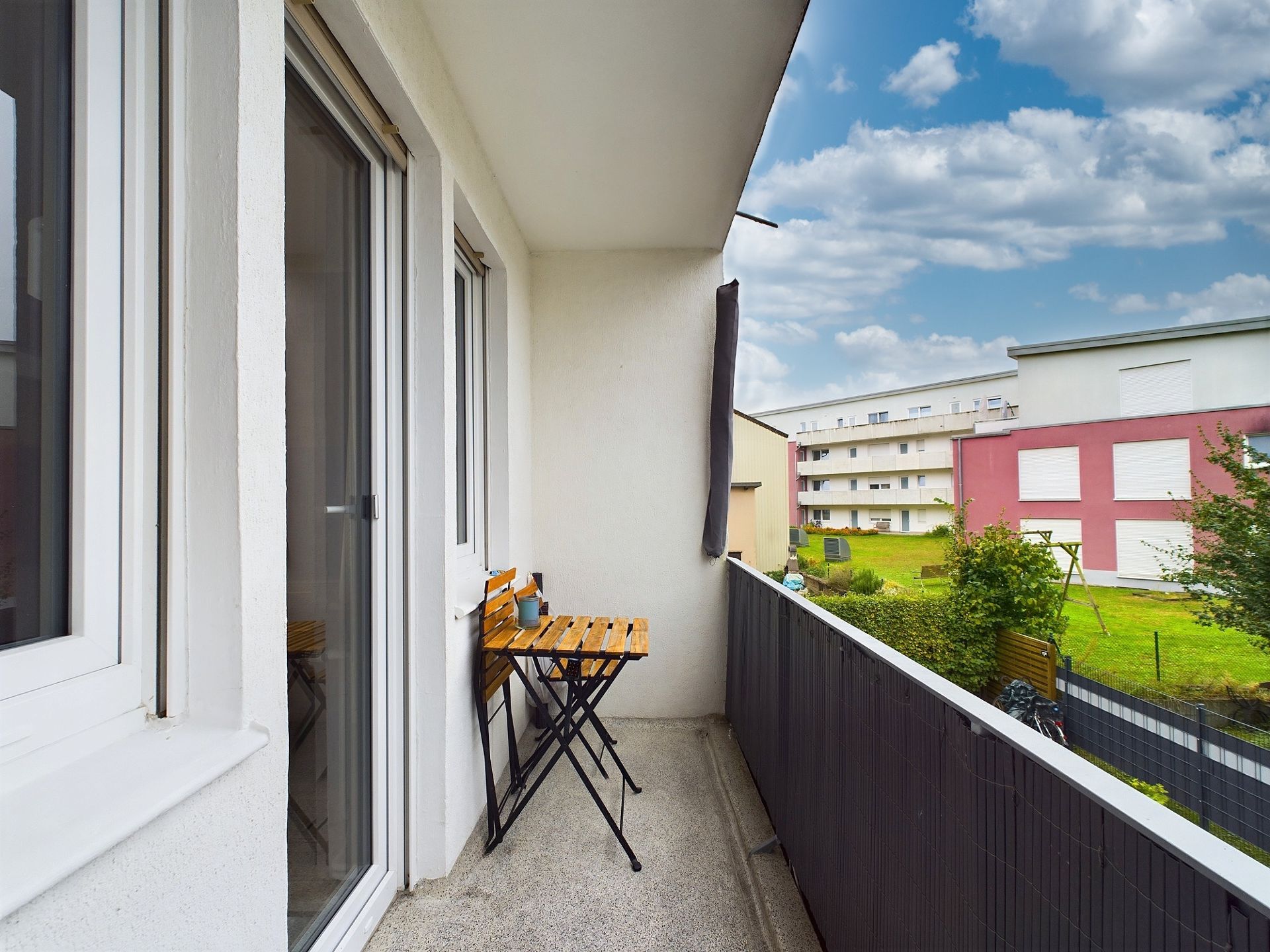 Schmaler Balkon mit kleinem Holztisch und zwei Stühlen, Blick auf Grünfläche und Wohngebäude.