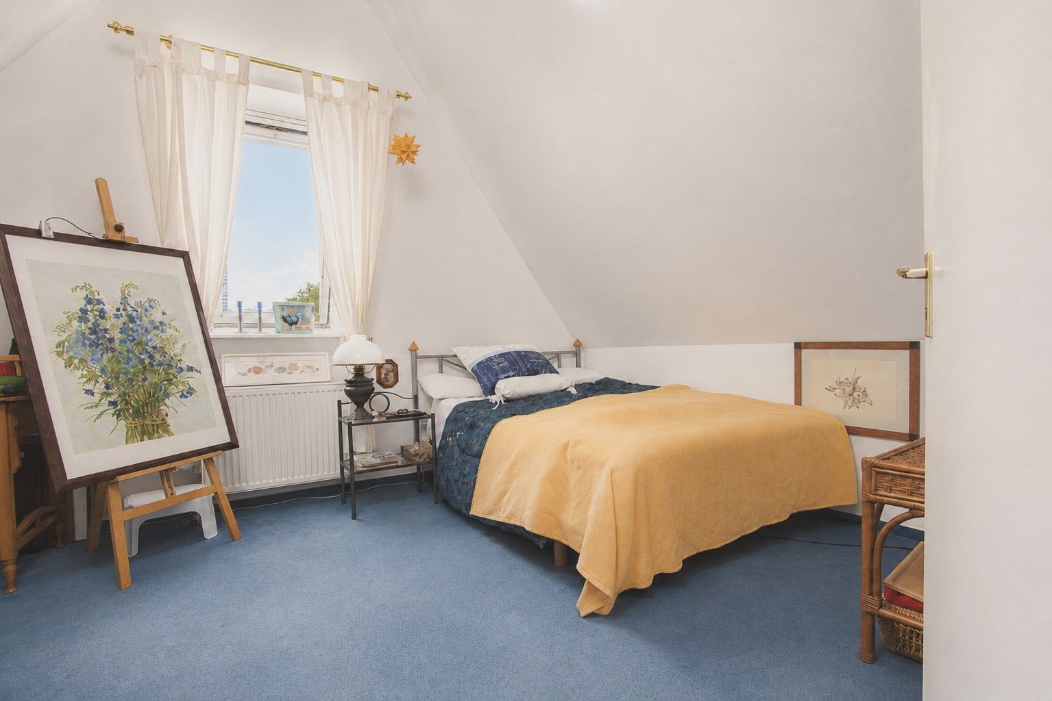 Helles Schlafzimmer mit Einzelbett, gelber Decke, blauem Teppich und Bildern an den Wänden.