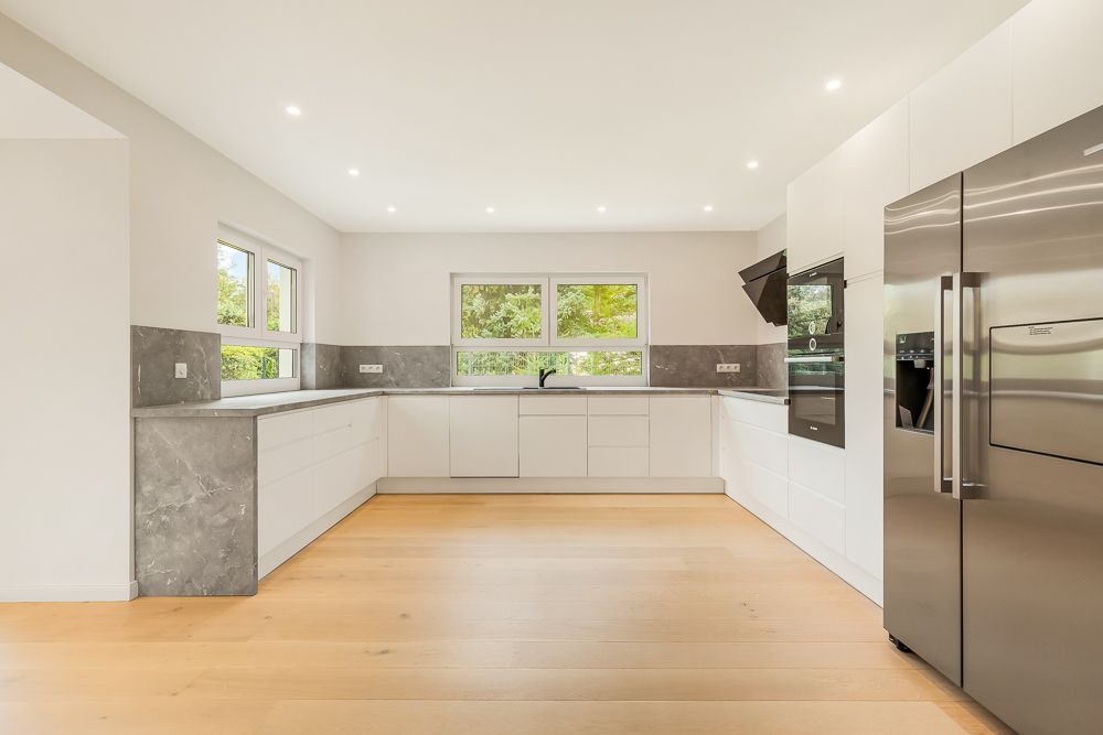 Moderne, helle Einbauküche mit großen Fenstern, weißen Schränken und Edelstahl-Kühlschrank.