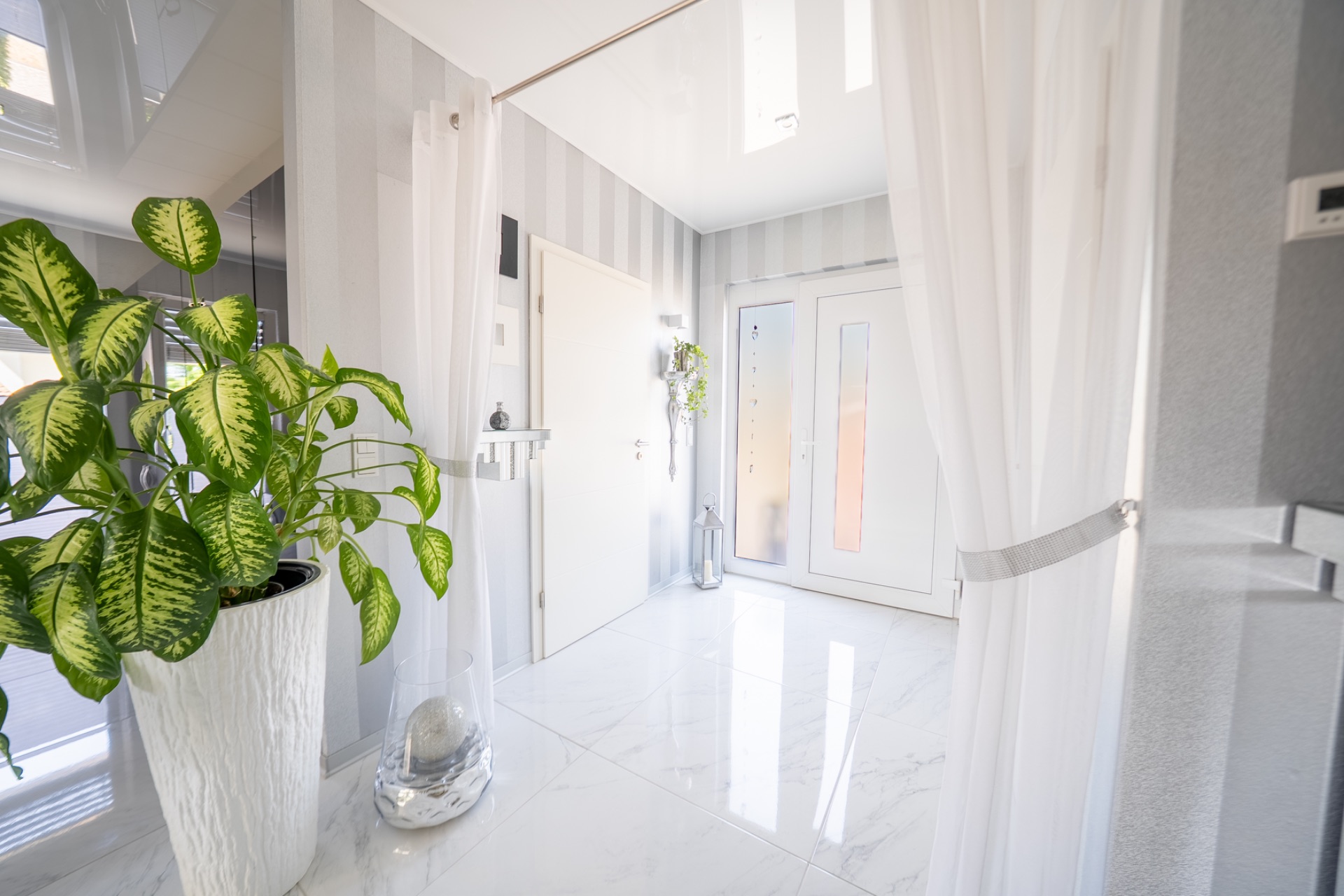 Heller Flur mit weißem Boden, weißen Türen, Vorhängen und einer großen grünen Pflanze im Blumentopf.