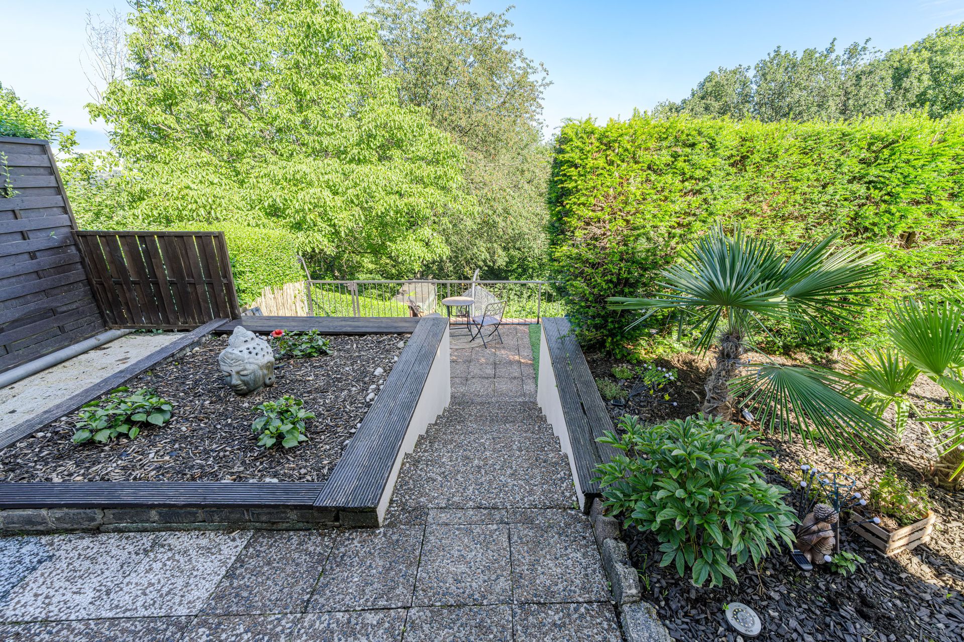 Garten mit Steintreppe, kleinen Pflanzen, einer Buddha-Statue und Sitzbereich mit Tisch und zwei Stühlen.