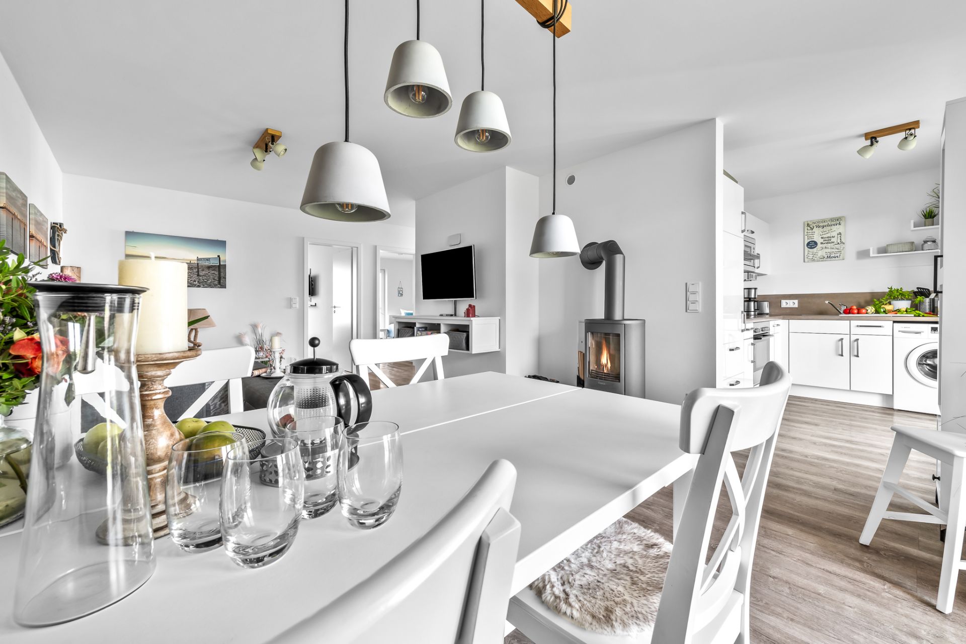 Helles Esszimmer mit weißem Tisch, Stühlen, Dekoration und Blick auf offene Küche mit Kaminofen.