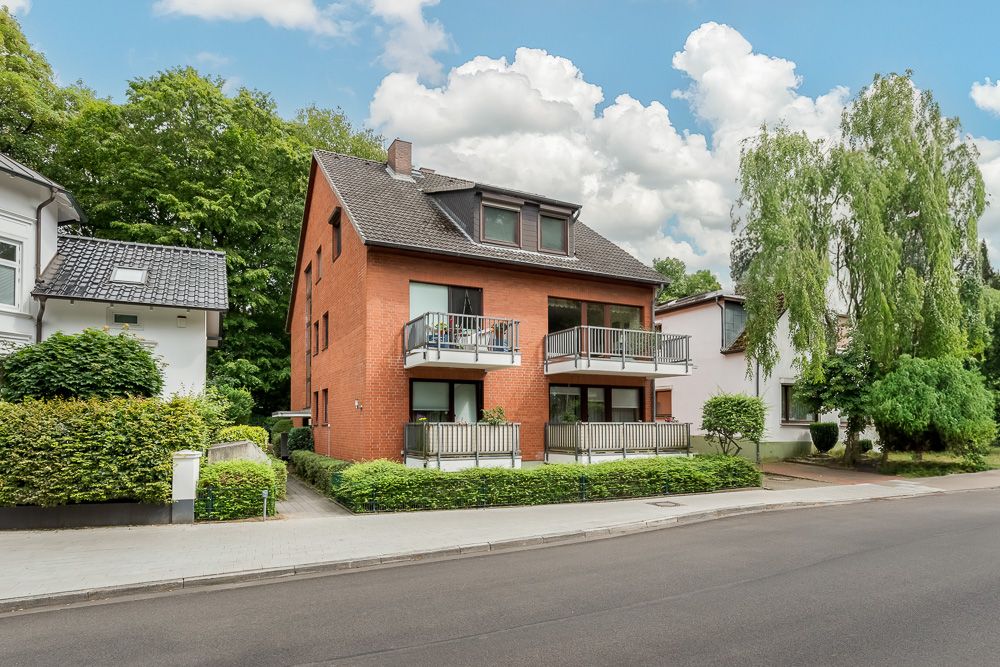 Modernes rotes Wohnhaus mit Balkonen, umgeben von Bäumen und Nachbarhäusern an einer ruhigen Straße.