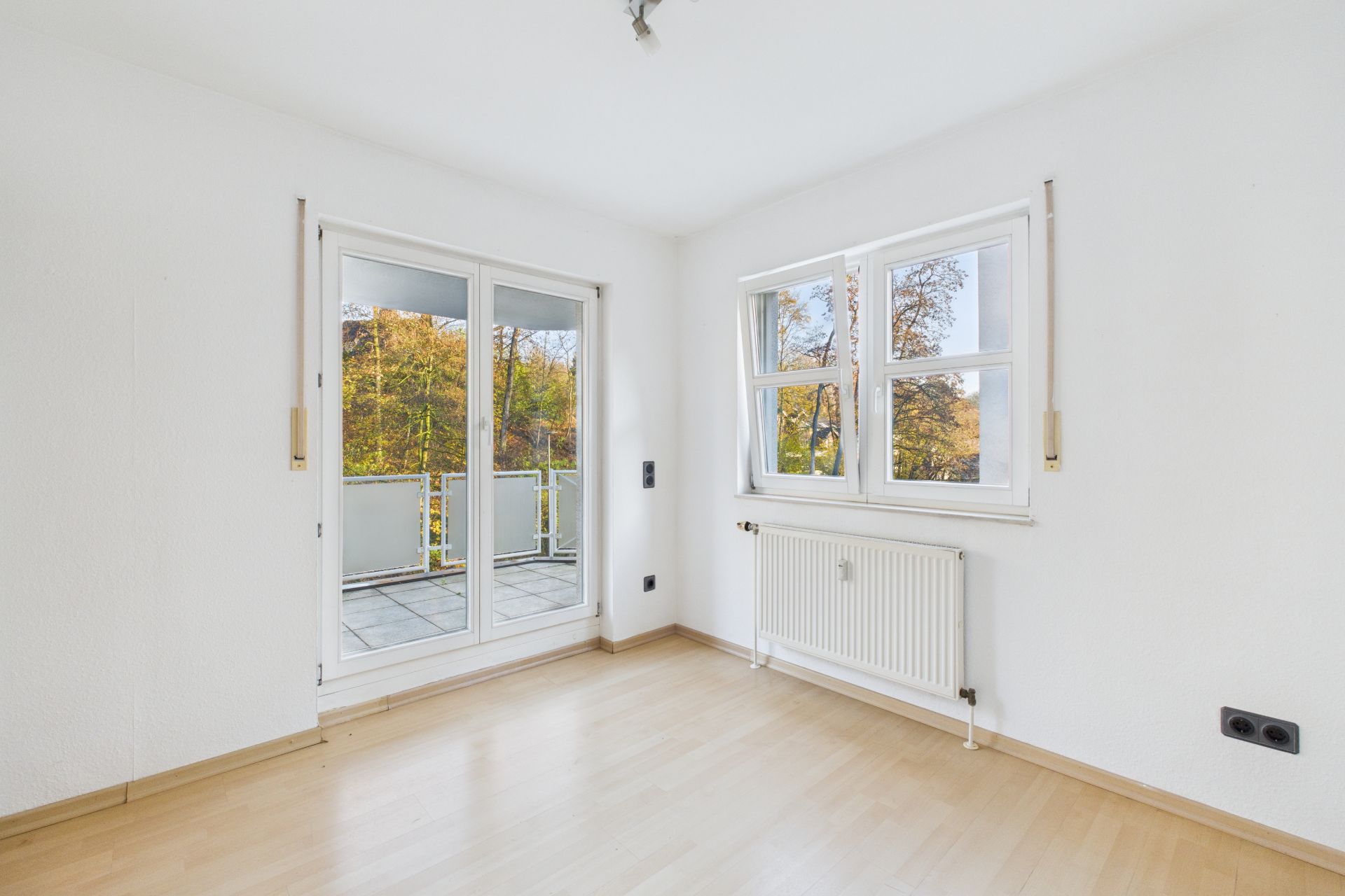 Helles Zimmer mit Holzfußboden, Fenster, Heizung und Zugang zu einem Balkon mit Blick ins Grüne.