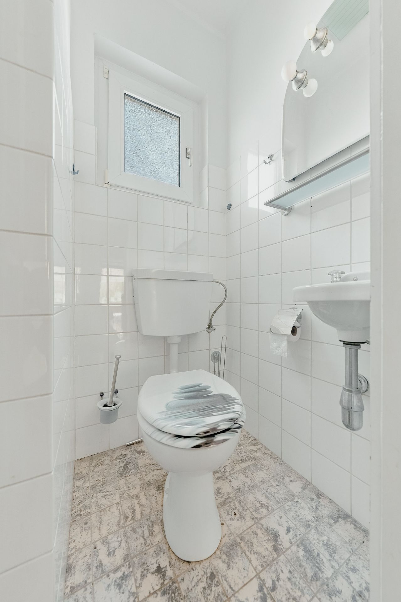 Kleines Badezimmer mit Toilette, Waschbecken, Spiegel und Fenster, weiße Fliesen an Wand und Boden.