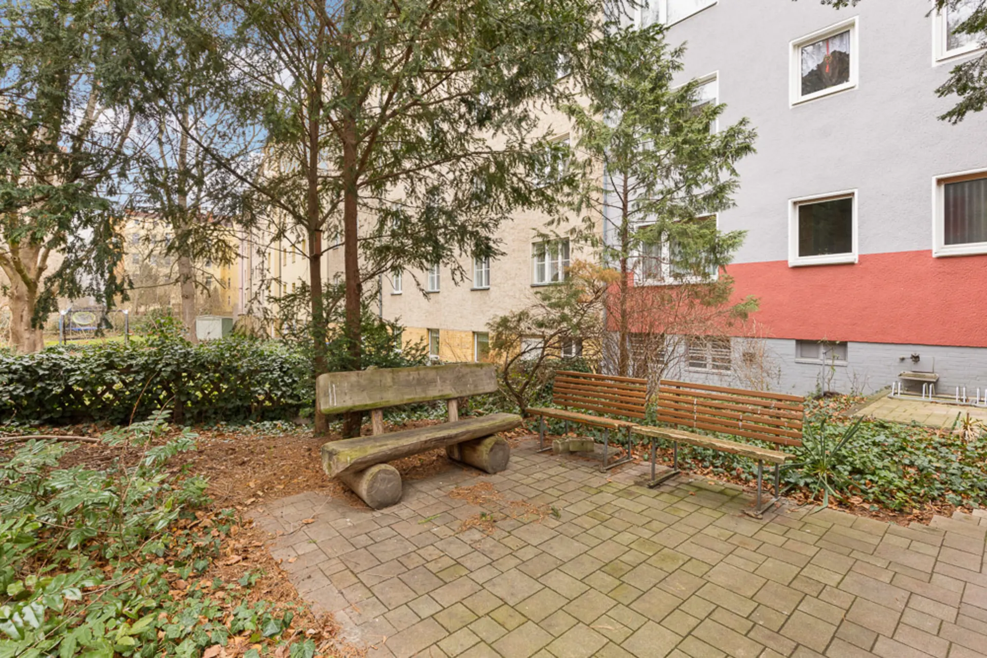 Gartenbereich mit zwei Holzbänken auf gepflastertem Boden, umgeben von Bäumen und Sträuchern.