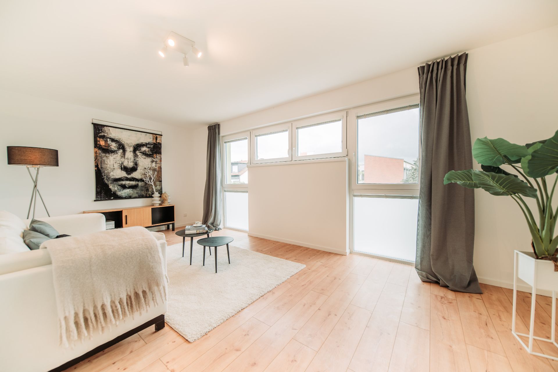Helles Wohnzimmer mit Sofa, Couchtischen, großem Fenster, Pflanze und Wandbild.