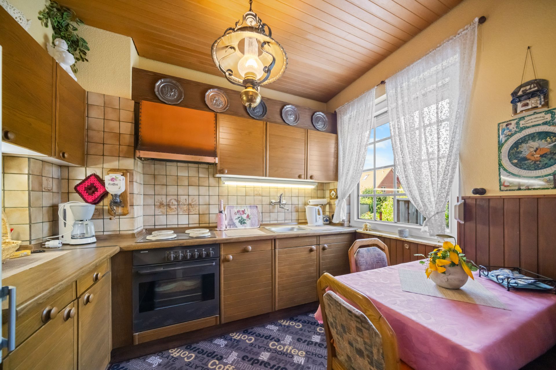 Gemütliche Küche mit Holzschränken, Esstisch, Herd, Kaffeemaschine und großem Fenster mit Vorhängen.