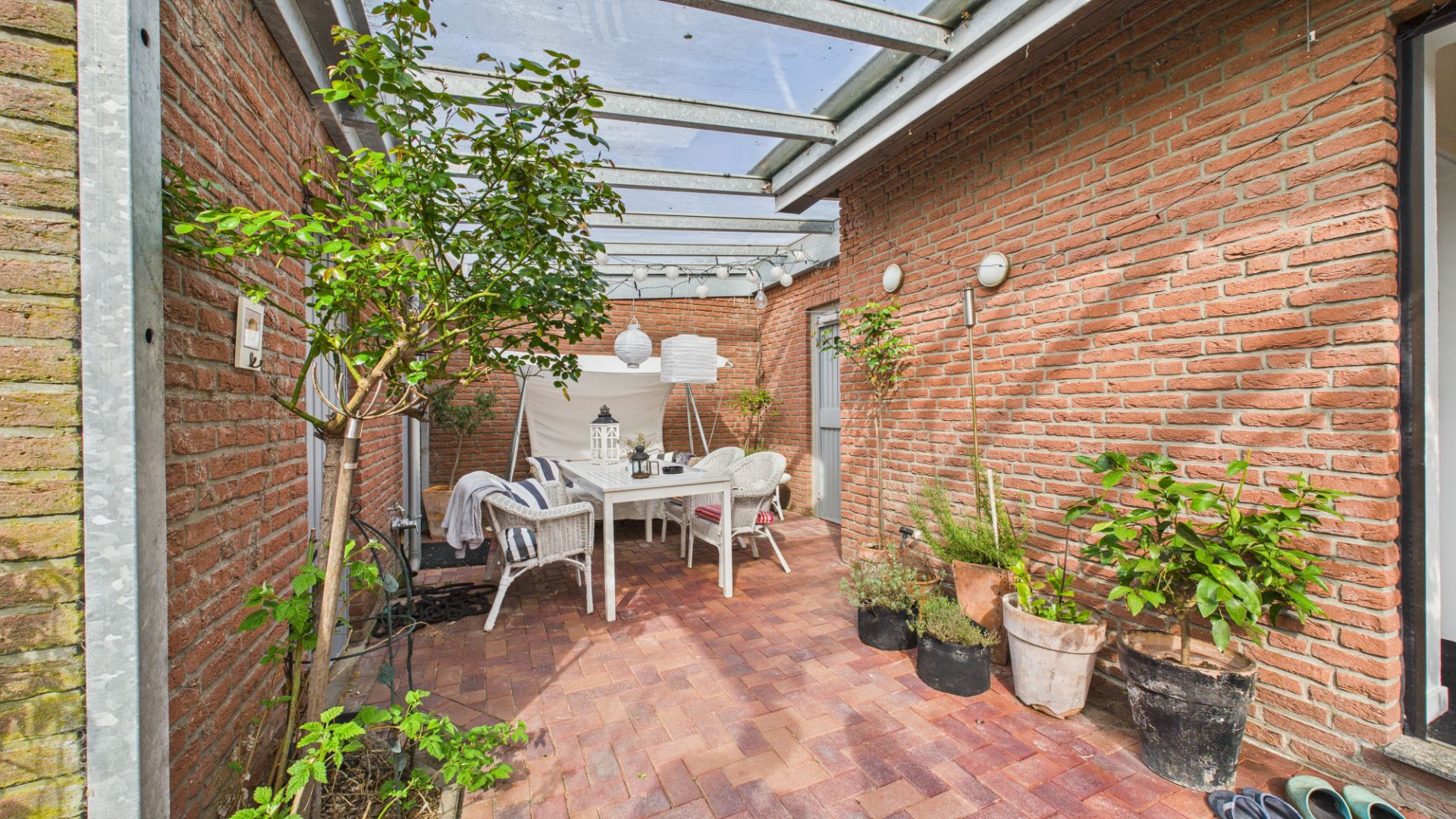Überdachte Terrasse mit Ziegelboden, Esstisch, Stühlen und mehreren Pflanzen in Töpfen.