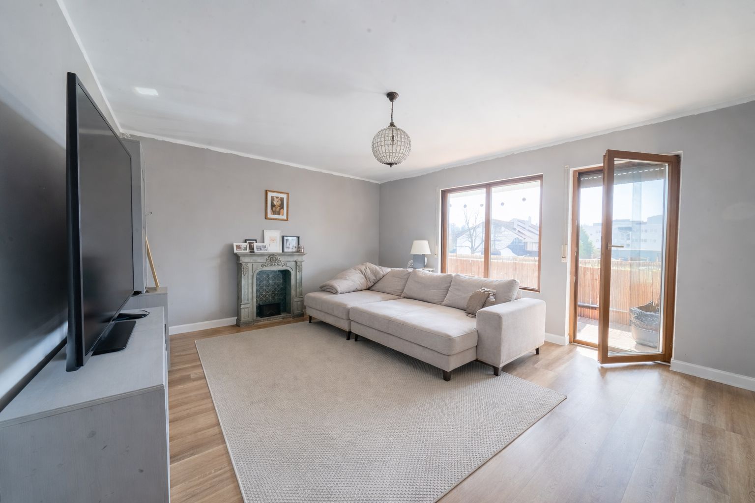 Helles Wohnzimmer mit großem Sofa, Teppich, Fernseher und offenem Zugang zum Balkon.
