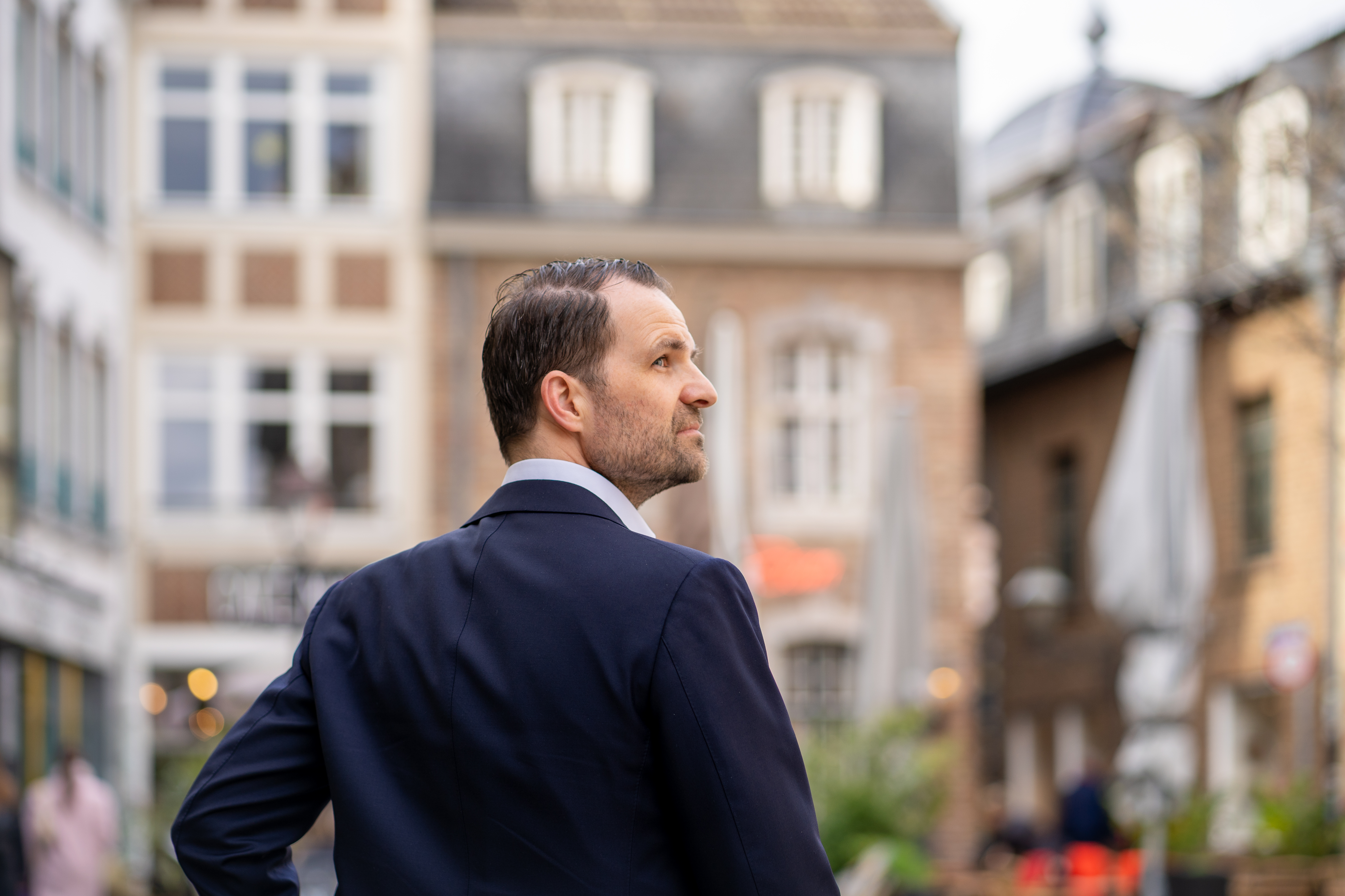 Ein Mann im Anzug steht vor mehreren Wohnhäusern in einer Stadt.