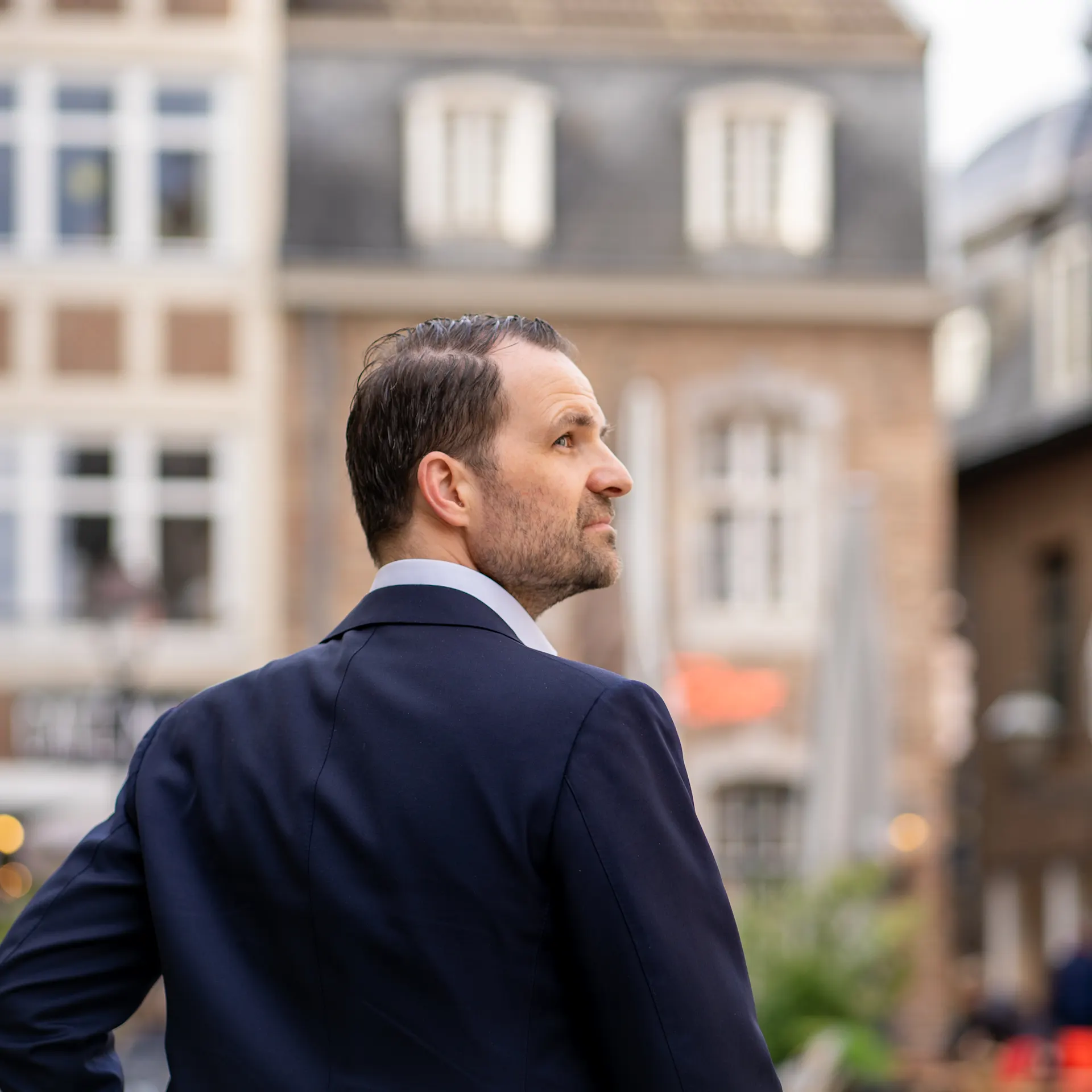 Ein Mann im Anzug steht vor mehreren Wohnhäusern in einer Stadt.