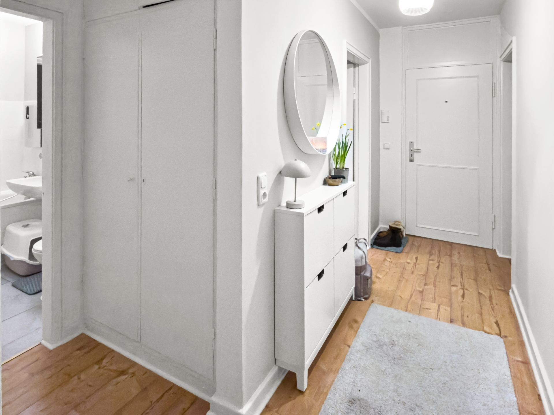 Heller Flur mit Holzfußboden, Schuhschrank, rundem Spiegel und Zugang zum Badezimmer.