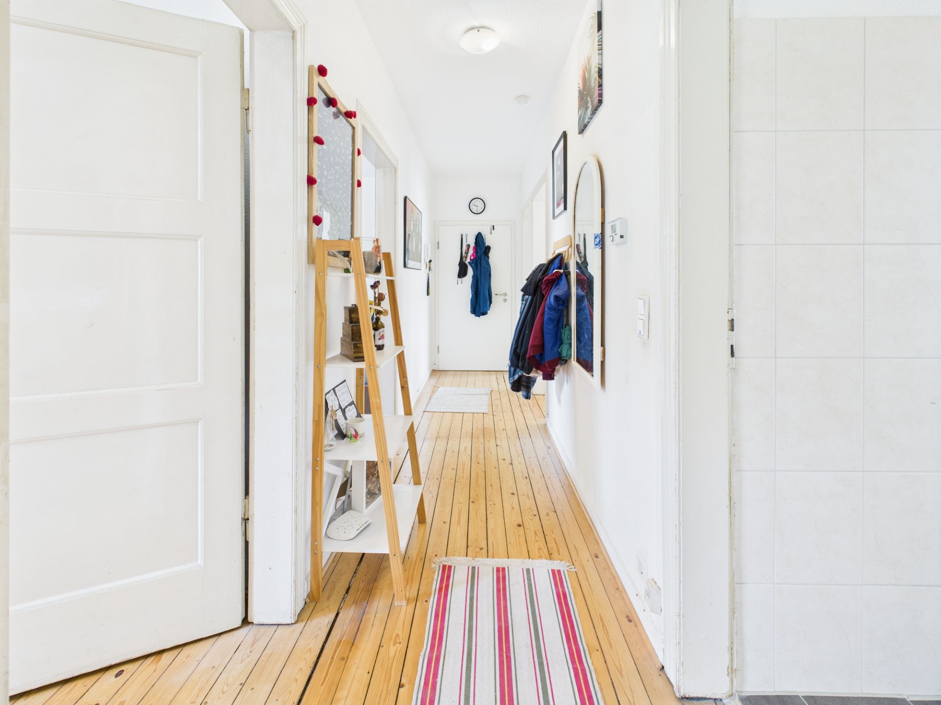 Heller Flur mit Holzfußboden, Teppichen, Regal, Wandbildern und Garderobe mit Jacken.