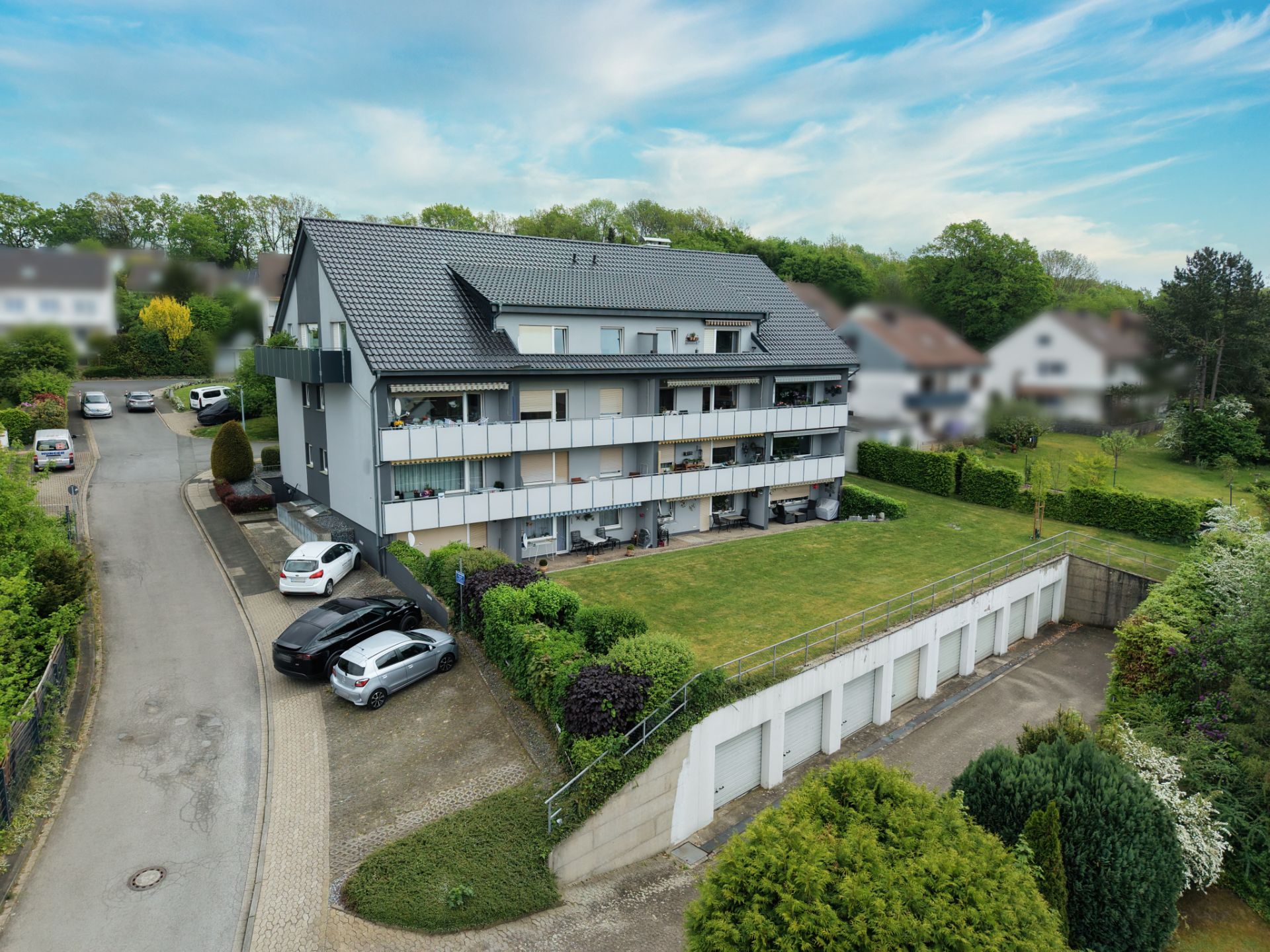 Mehrfamilienhaus mit Balkonen, Rasenfläche, Garagen und parkenden Autos auf einer ruhigen Wohnstraße.