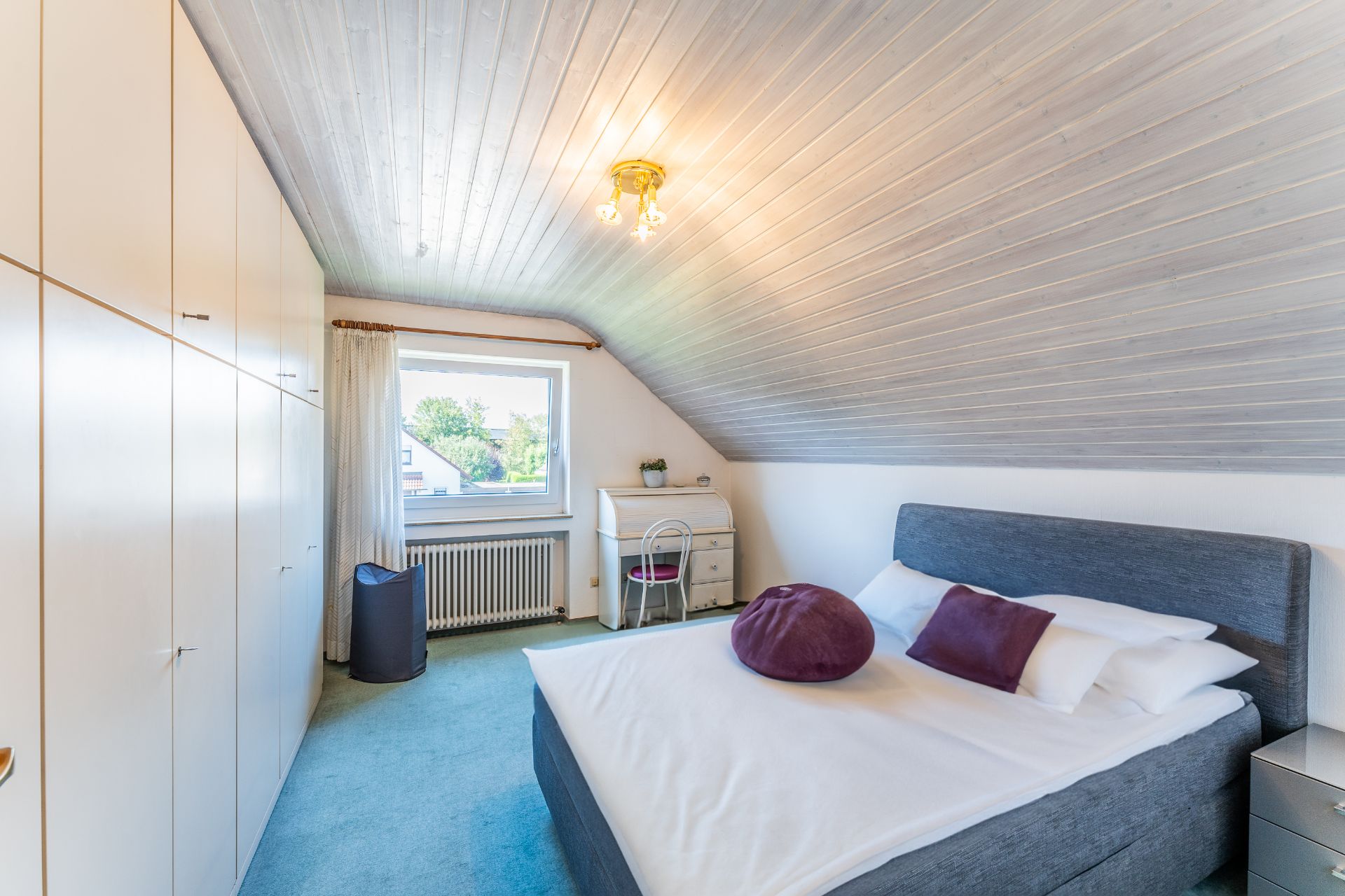 Helles Schlafzimmer mit großem Bett, Einbauschränken und Schreibtisch unter einem Fenster.