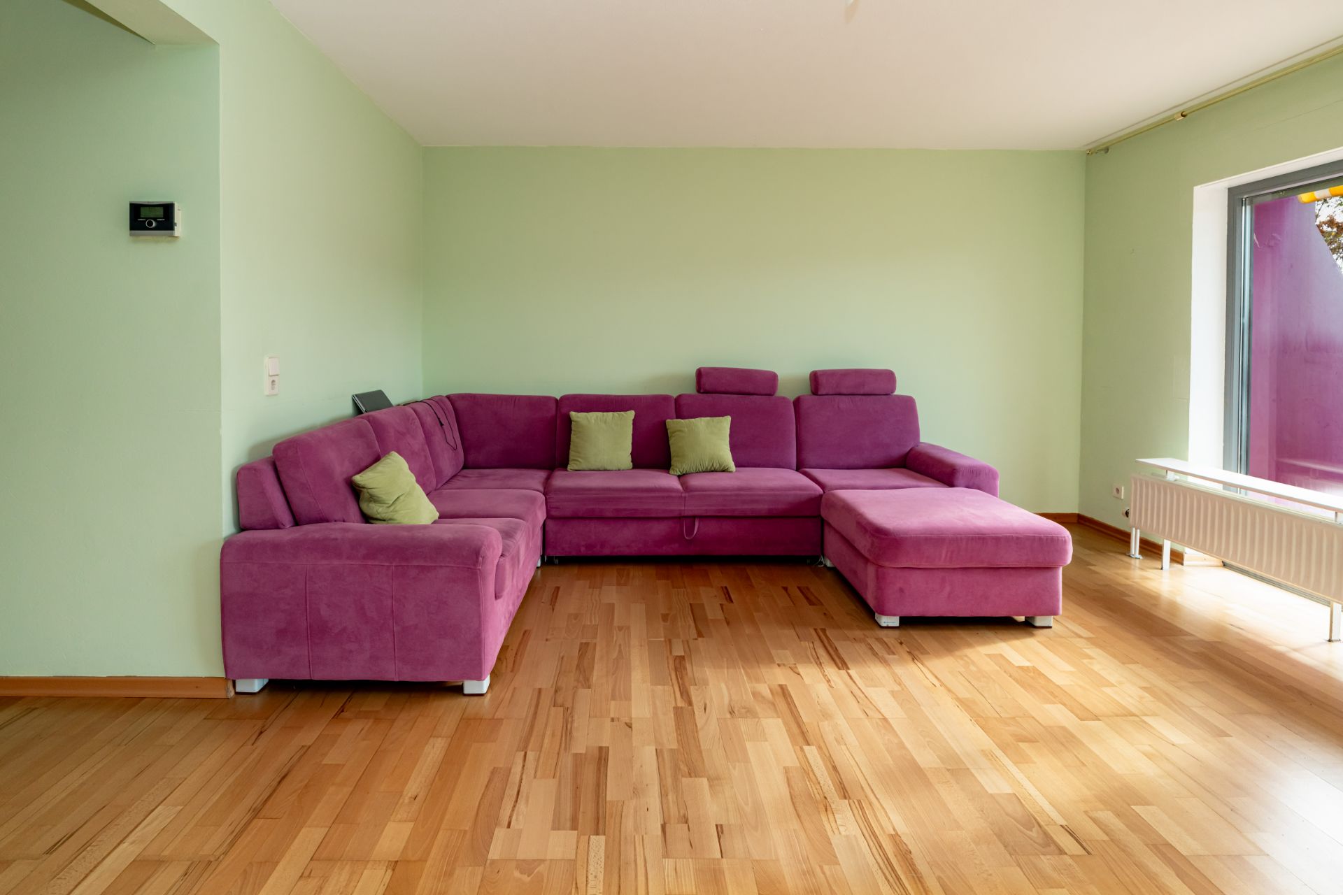 Wohnzimmer mit lila Ecksofa und grünen Kissen auf hellem Holzboden.