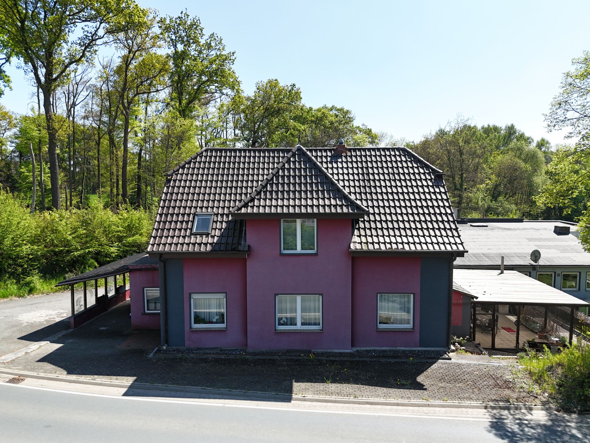 Lila Wohnhaus mit dunklem Dach und Carport, umgeben von Bäumen.