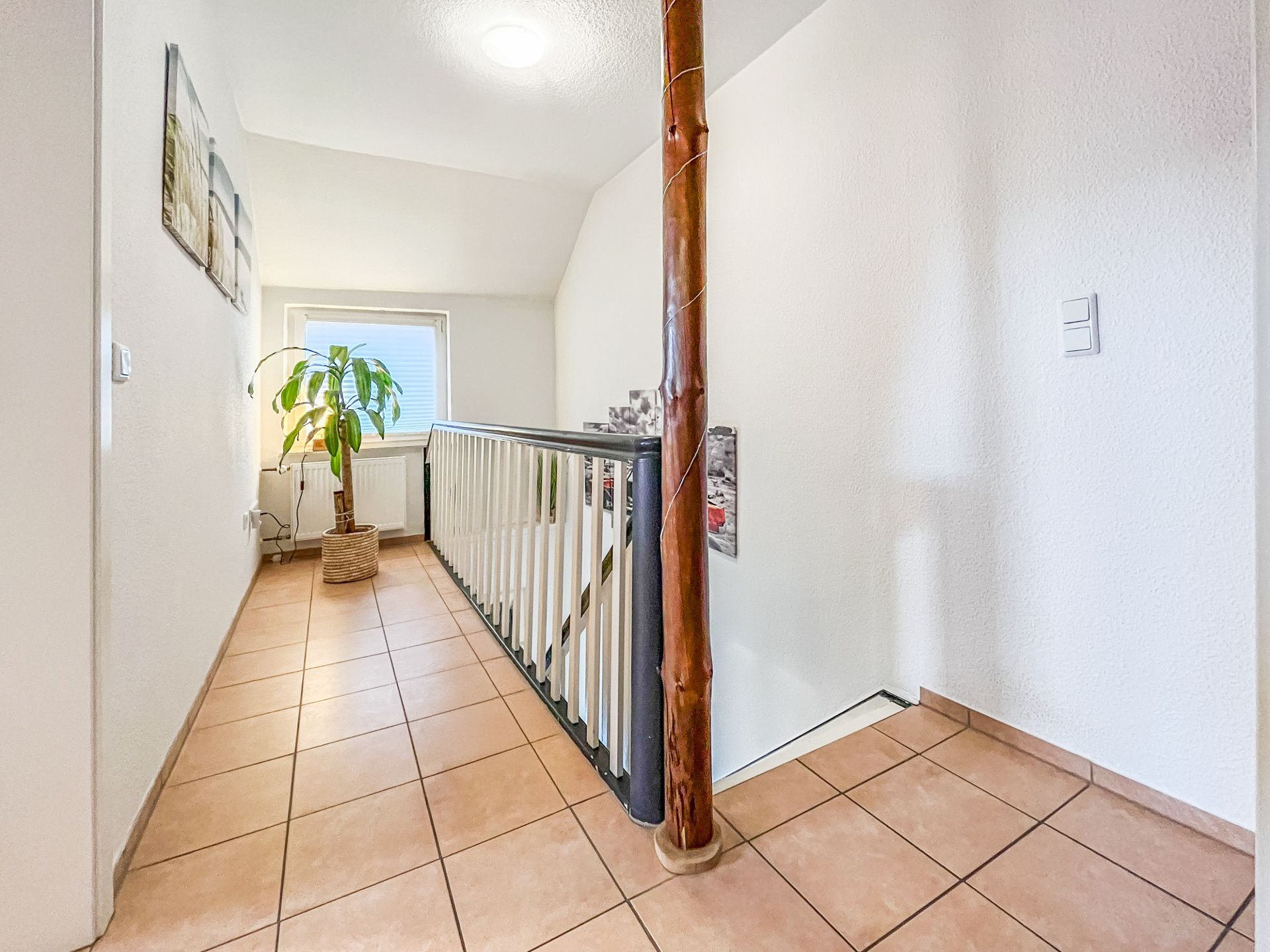 Heller Flur mit Fliesenboden, Pflanze im Topf und Geländer bei einer Treppe.