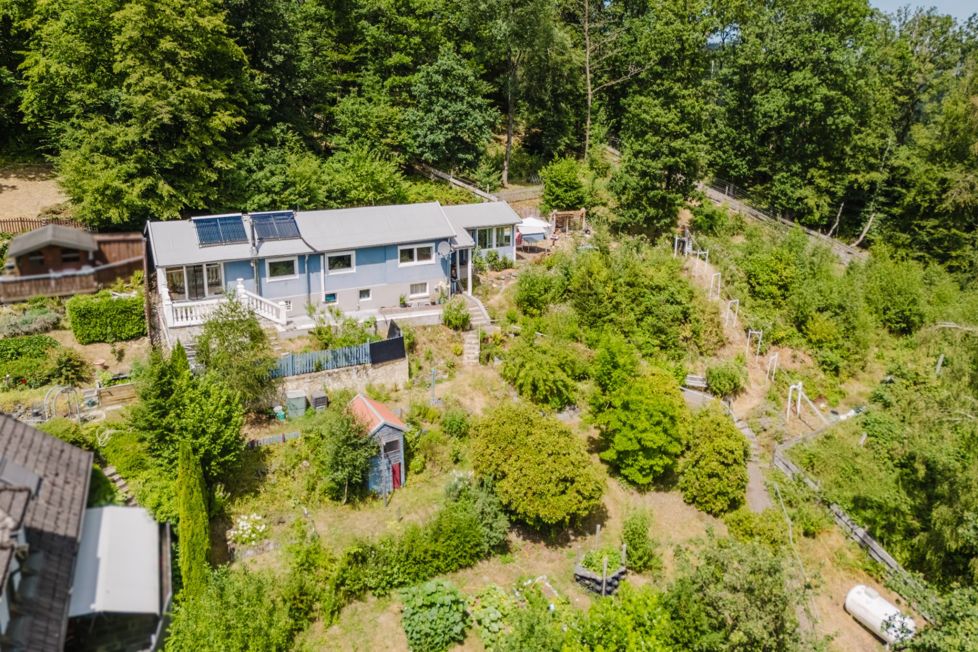 Langes, blaues Wohnhaus mit Solarpanels im Grünen, umgeben von Garten und Bäumen.