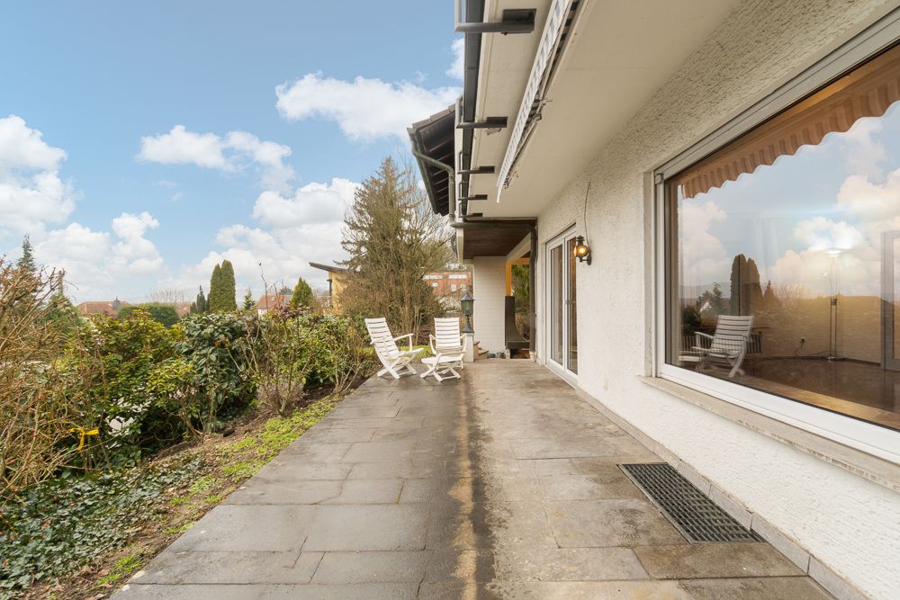 Große Terrasse mit weißen Gartenstühlen neben einem Haus, umgeben von Büschen und Pflanzen.