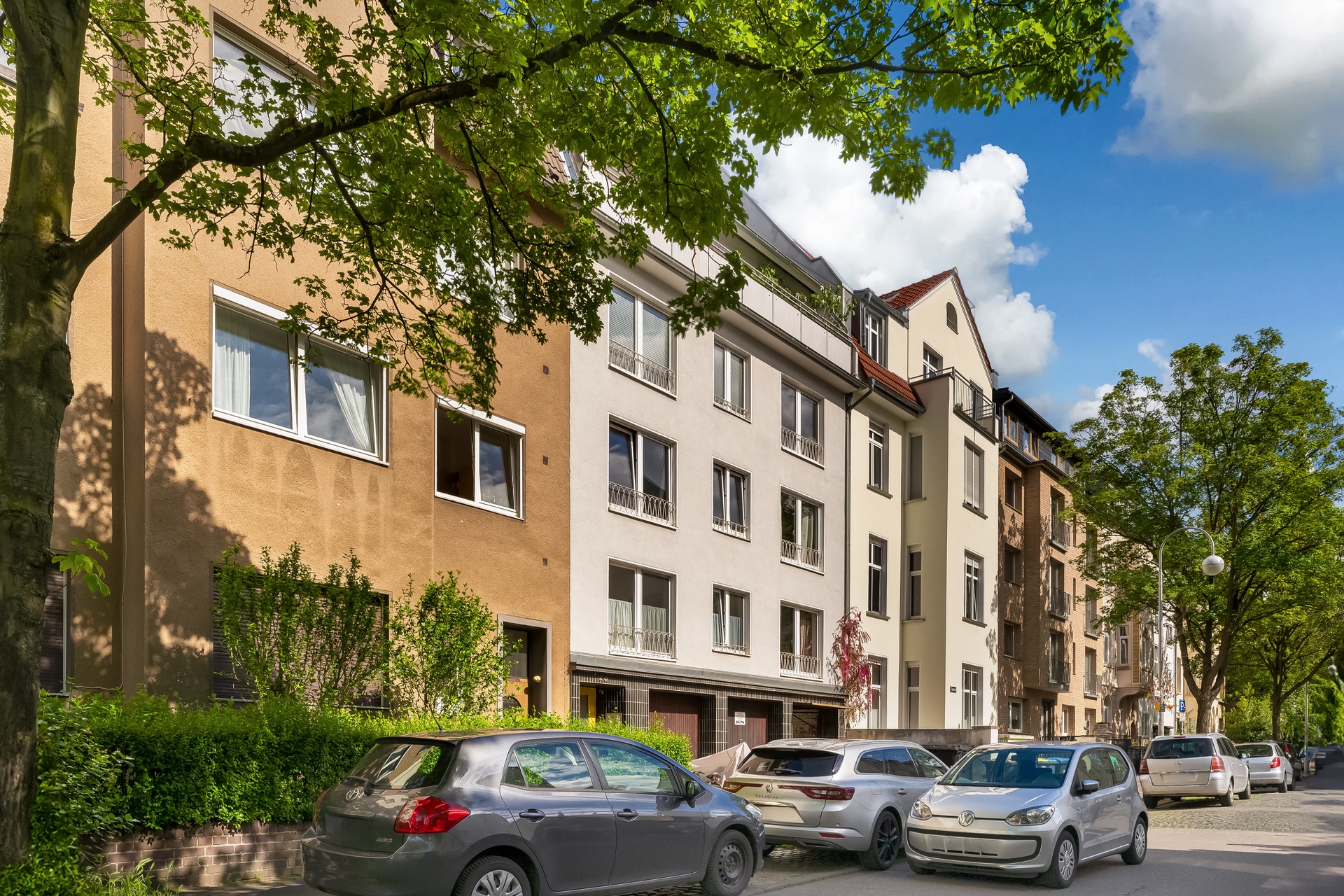 Mehrstöckige Wohnhäuser mit geparkten Autos an einer baumbestandenen Straße.