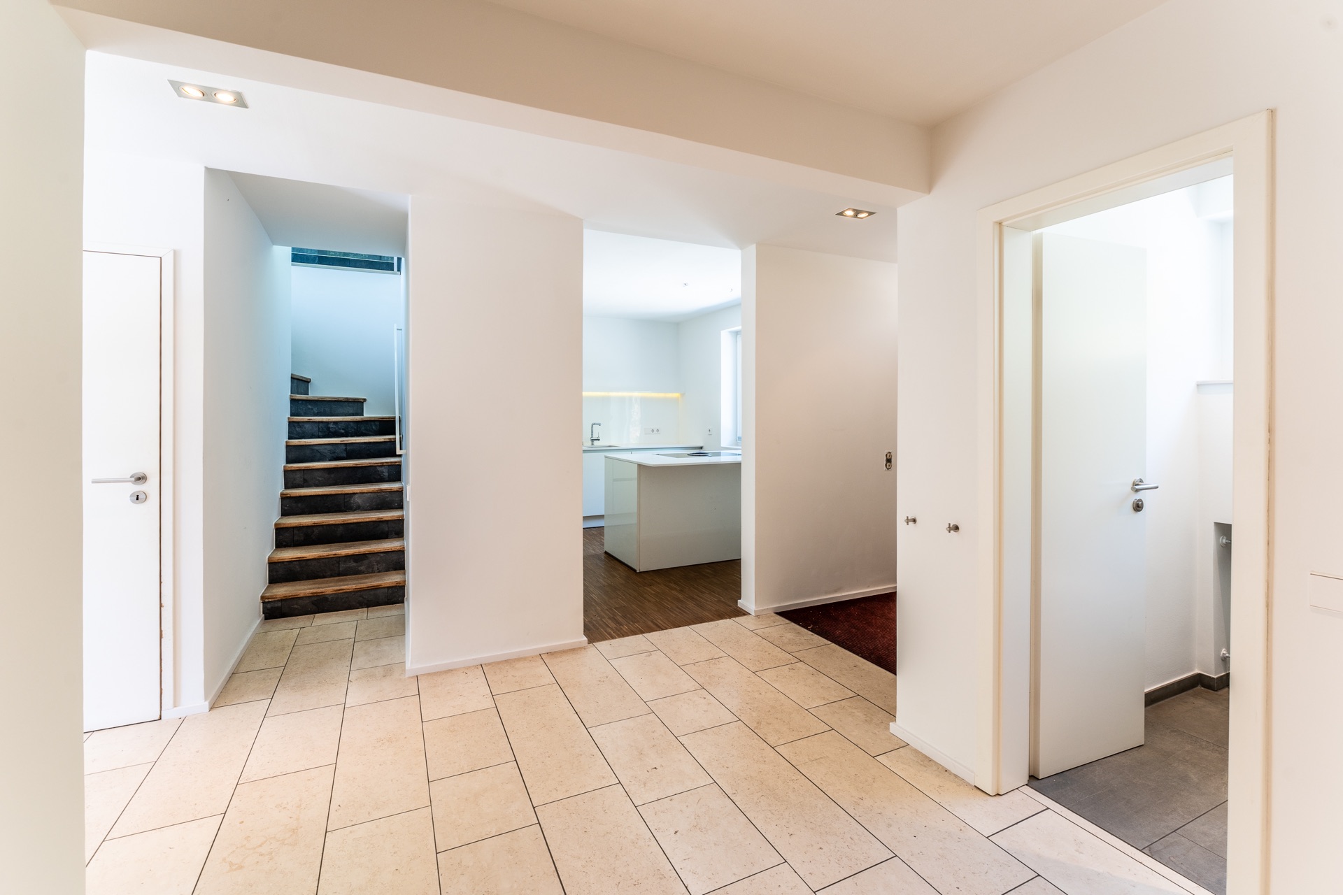 Heller Flur mit Fliesenboden, Treppe, Tür und Blick in eine moderne Küche.
