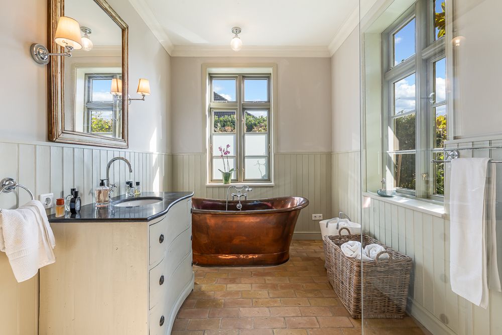 Helles Badezimmer mit freistehender Kupferbadewanne, großem Spiegel und zwei Fenstern.