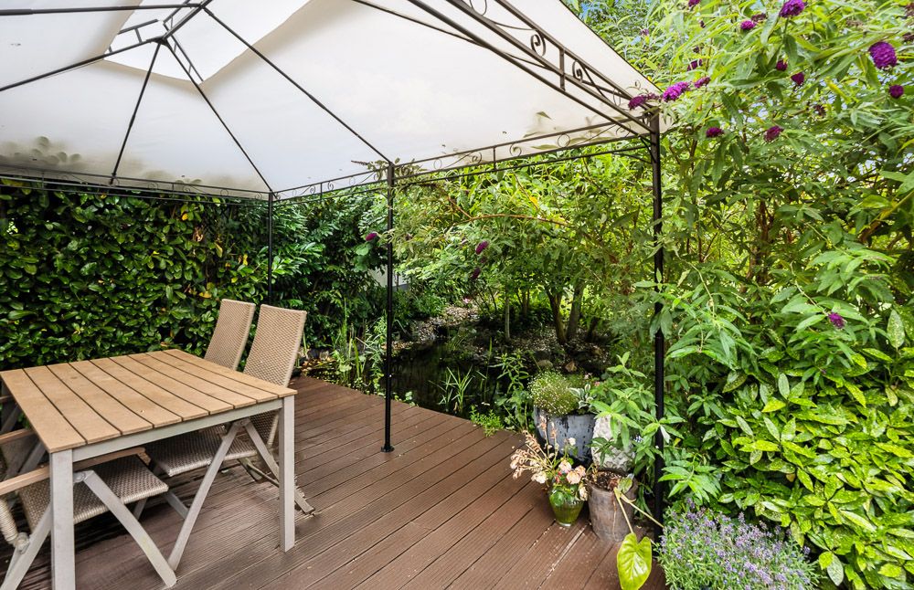 Überdachte Terrasse mit Holztisch und Stühlen, umgeben von dichtem grünen Pflanzenwuchs.
