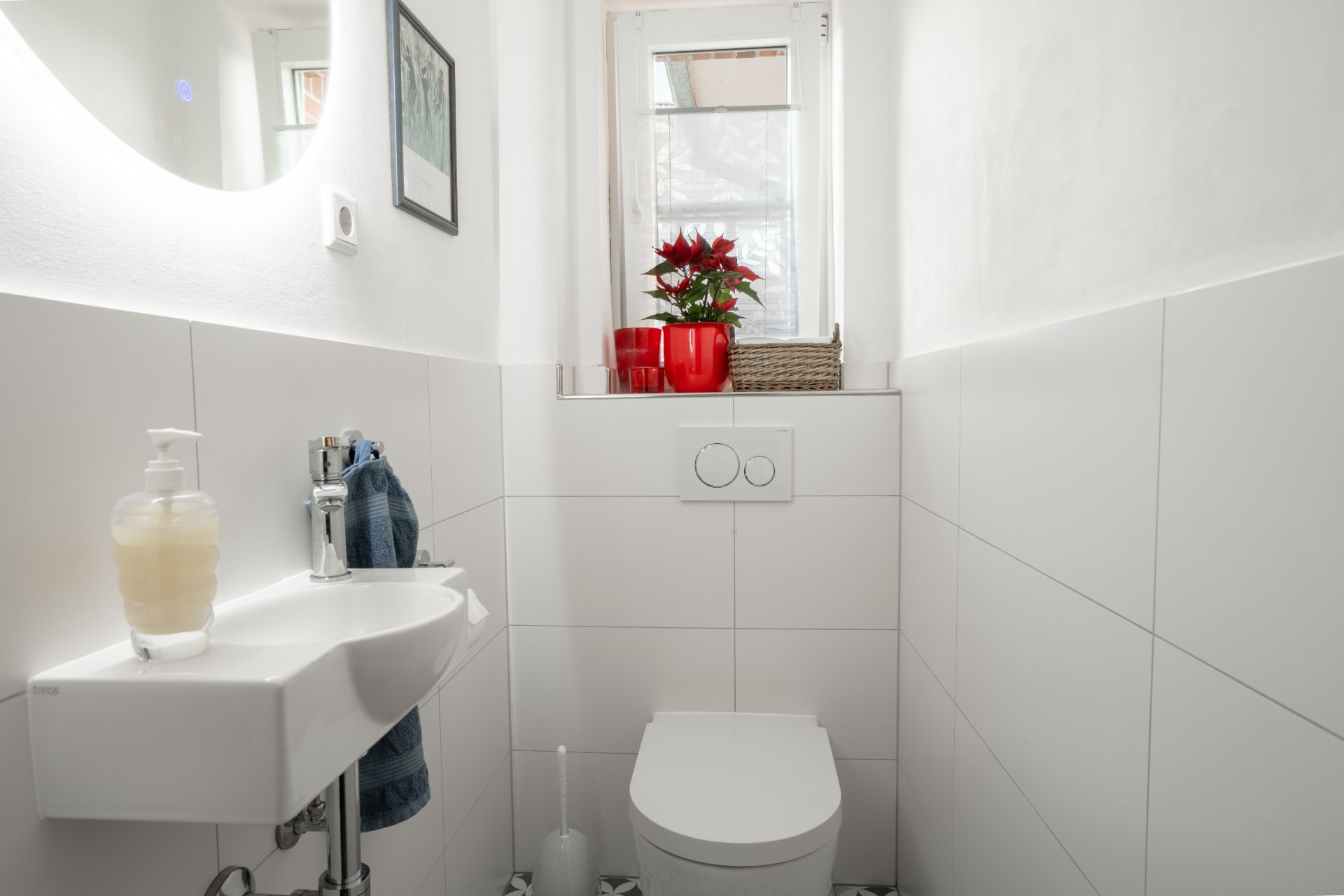 Kleines, weiß gefliestes Badezimmer mit Toilette, Waschbecken und Fenster mit Blumen in roten Töpfen.