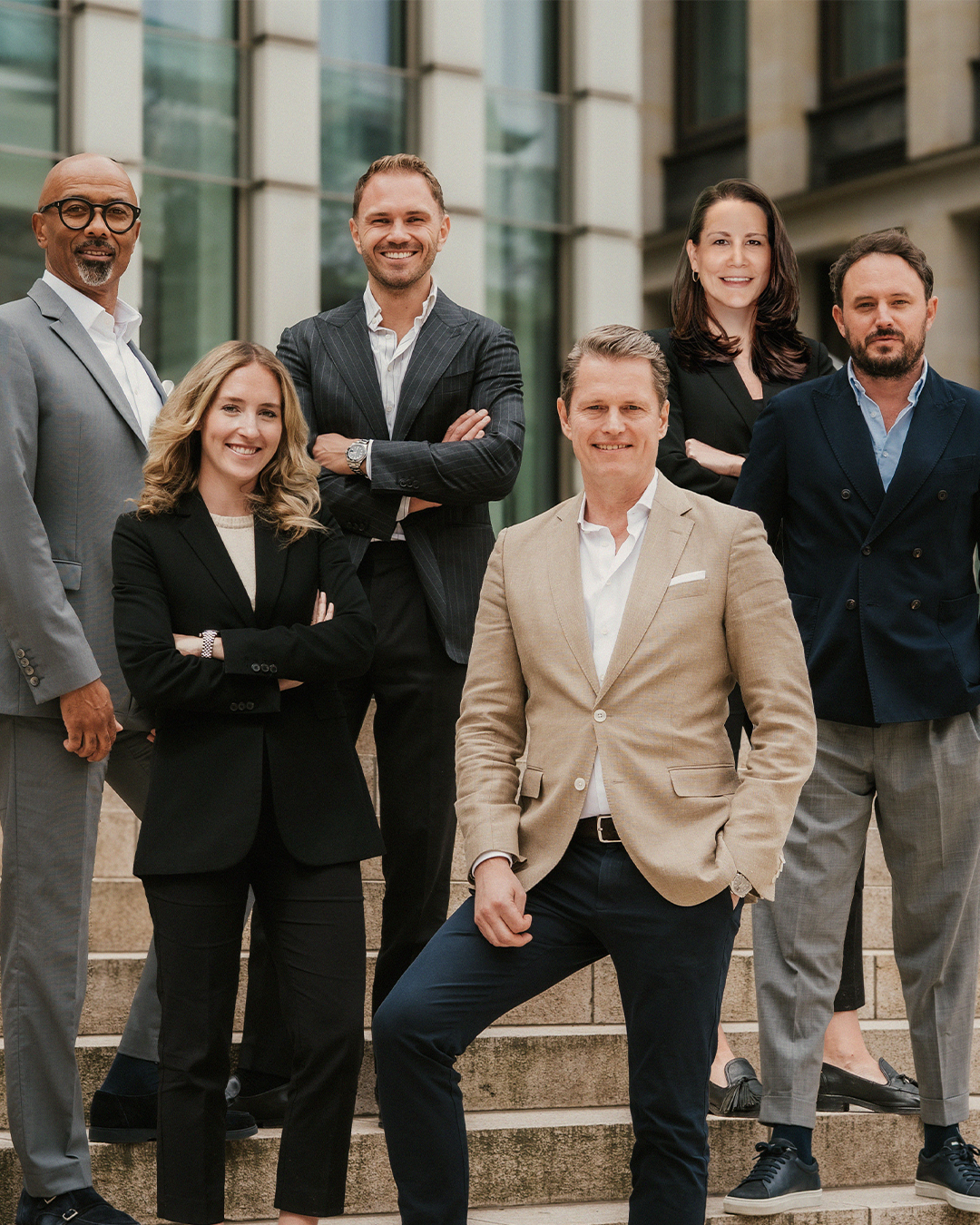 Sechs Personen in Anzügen stehen auf steinernen Außentreppen vor einem modernen Gebäude.