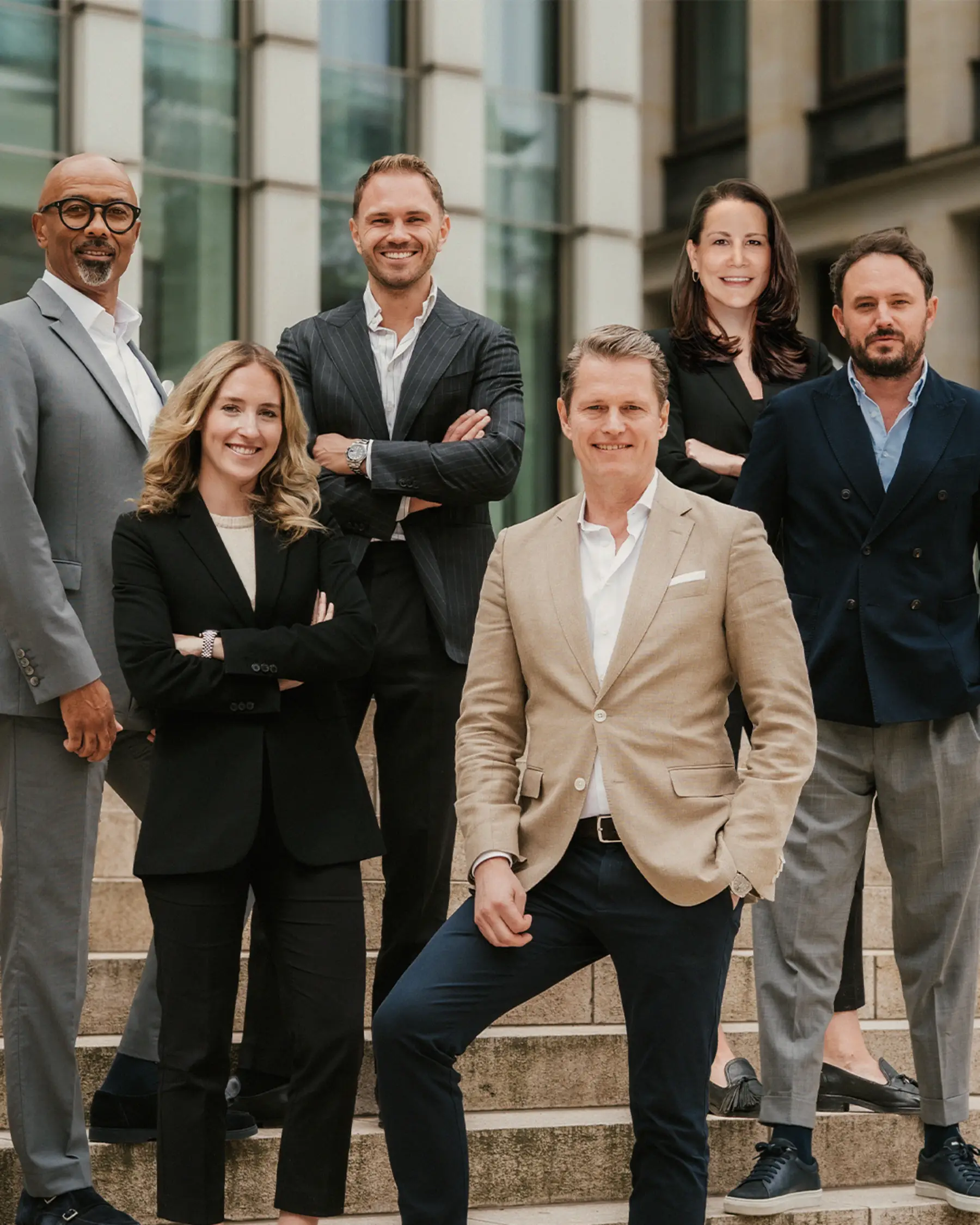 Sechs Personen in Anzügen stehen auf steinernen Außentreppen vor einem modernen Gebäude.