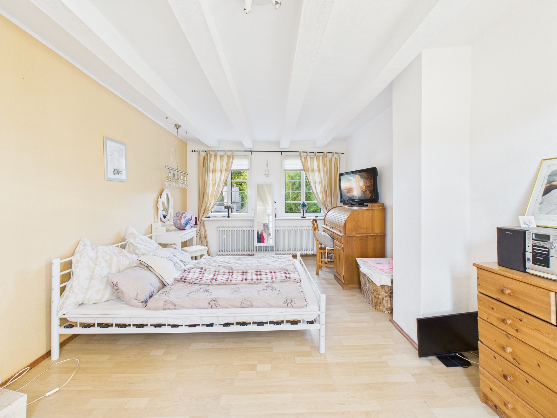 Helles Schlafzimmer mit Doppelbett, Holzschrank, Schreibtisch, Fernseher und zwei Fenstern mit Vorhängen.