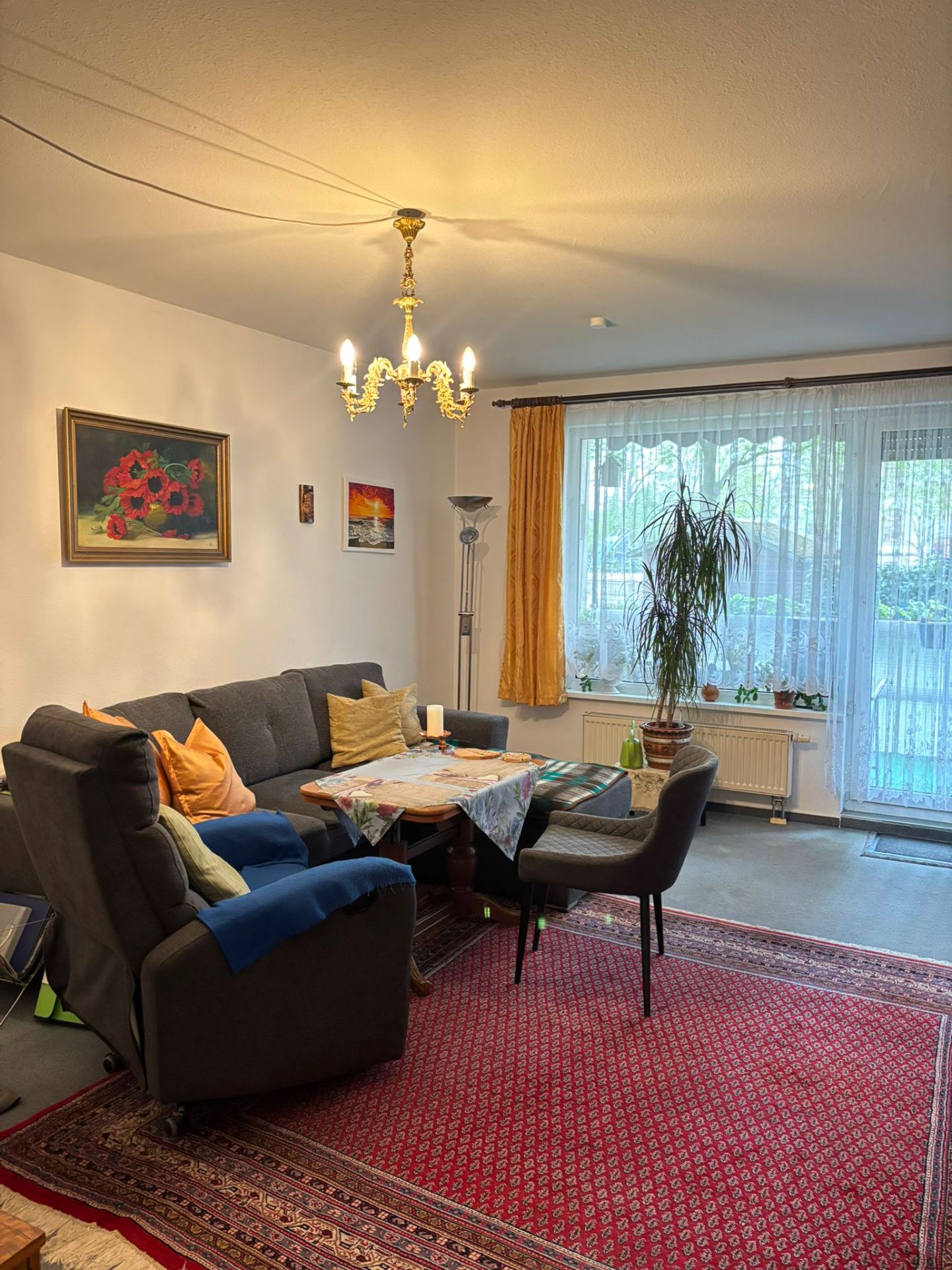 Wohnzimmer mit Ecksofa, Teppich, Couchtisch, Pflanze am Fenster und Kronleuchter an der Decke.