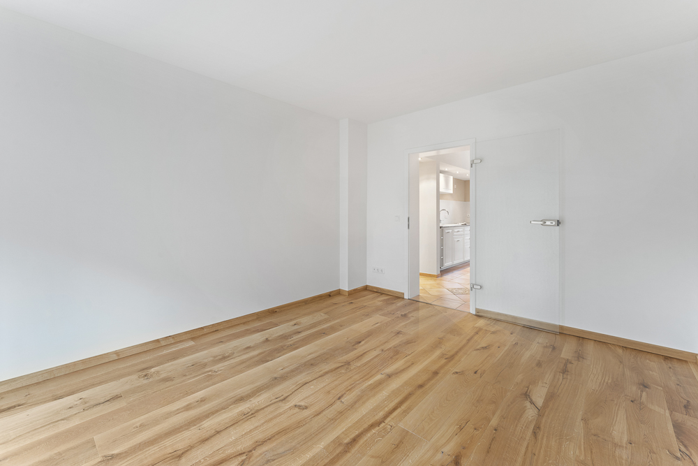 Leeres Zimmer mit Holzfußboden und Glastür, Blick in eine helle Küche.