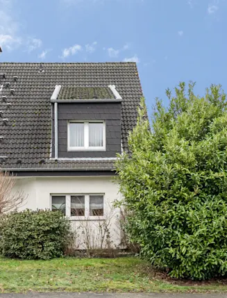 Vorderansicht eines Einfamilienhauses mit großem Busch und kurzem Baum im Vorgarten.