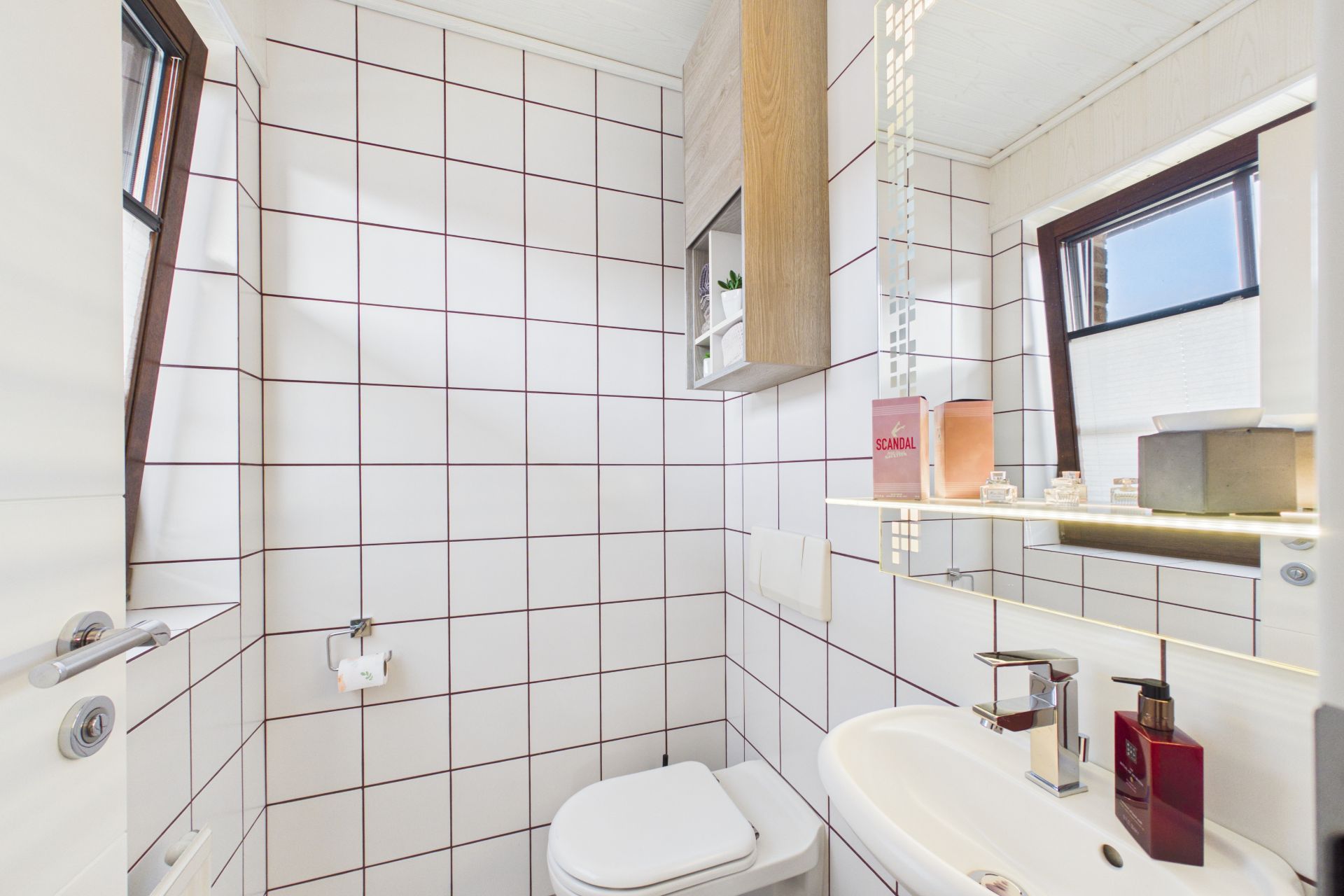 Kleines Bad mit weiß gefliesten Wänden, WC, Waschbecken, Spiegel und Fenster.