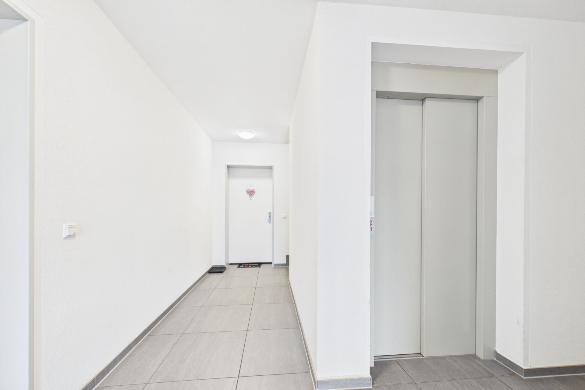 Heller Flur mit Fliesenboden, Aufzug rechts und Wohnungstür am Ende des Gangs.