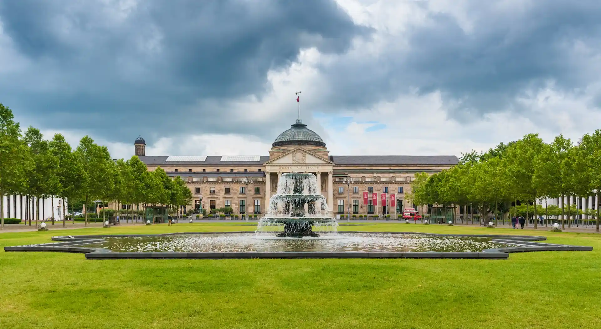 Großer Springbrunnen auf einer Wiese mit symmetrisch gepflanzten Bäumen und einem großen Gebäude im Hintergrund.