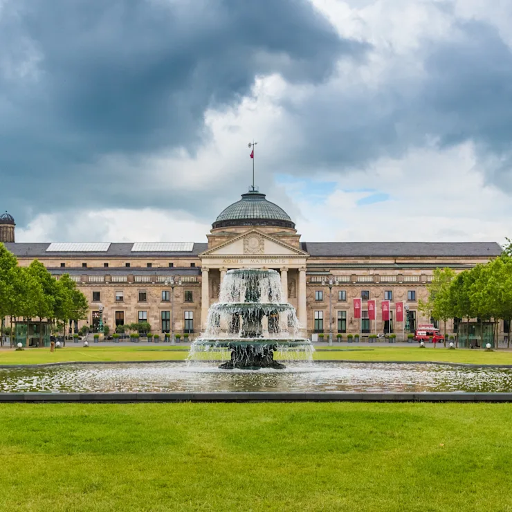 Großer Springbrunnen auf einer Wiese mit symmetrisch gepflanzten Bäumen und einem großen Gebäude im Hintergrund.