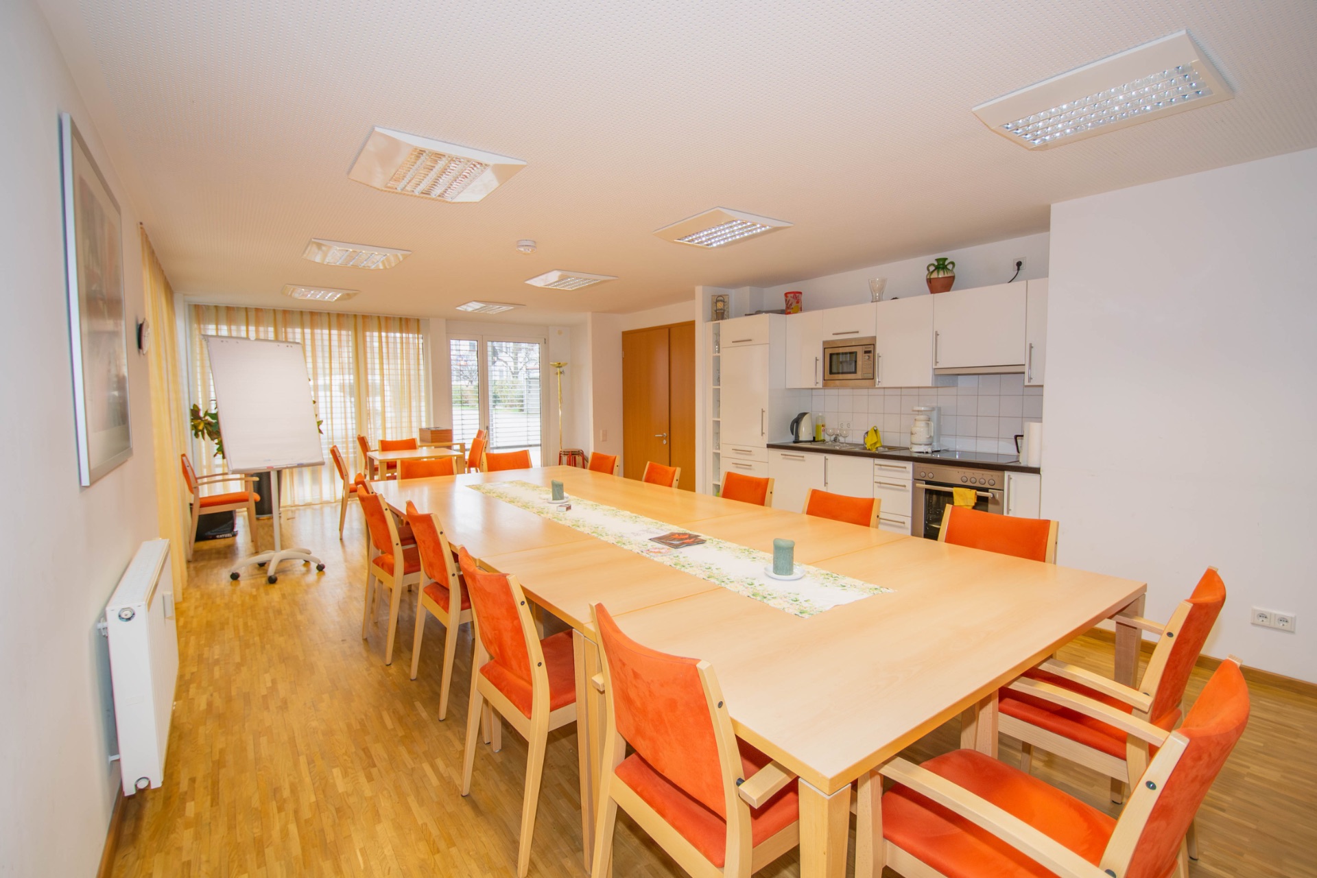 Heller Raum mit großem Holztisch, orange Stühlen, Küchenzeile und Flipchart.