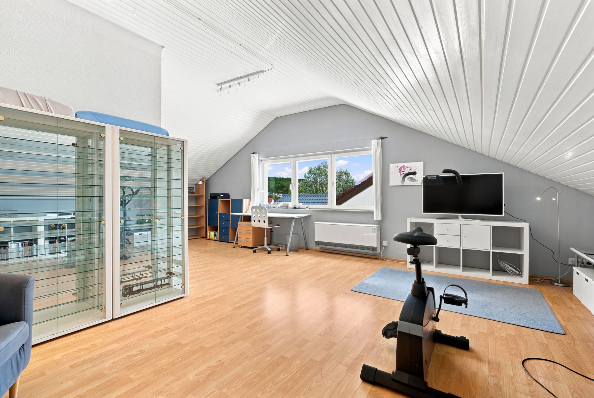 Helles Dachzimmer mit Schreibtisch, Glasvitrine, Heimtrainer und Fernseher auf Holzfußboden.