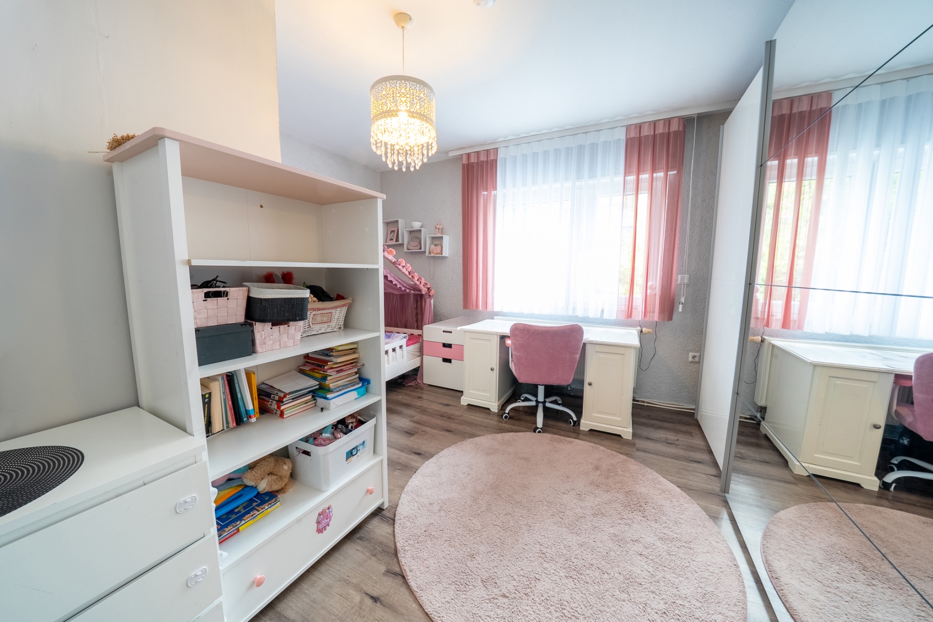 Kinderzimmer mit Schreibtisch, Regal, pinkem Teppich und großem Fenster mit Vorhängen.