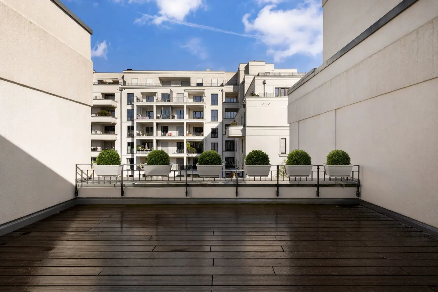 Große Terrasse mit Holzbelag, fünf Pflanzkübel und Blick auf ein Wohngebäude.